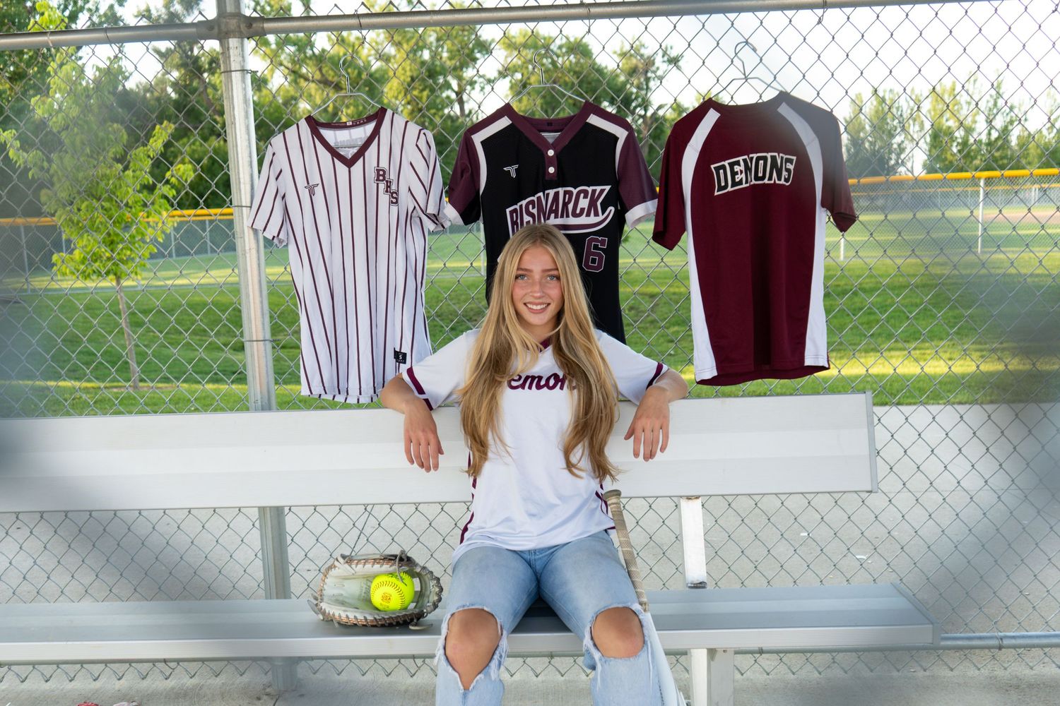 Smiling teenage girl sitting on bleachers with softball jerseys displayed on fence behind her.