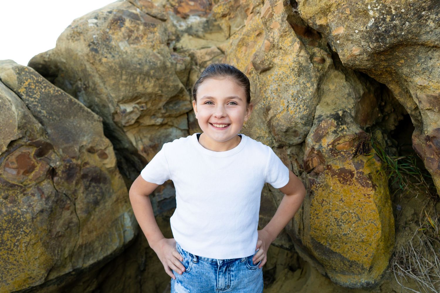 Smiling young girl in white t-shirt and jeans posing with hands on hips near large rocks.