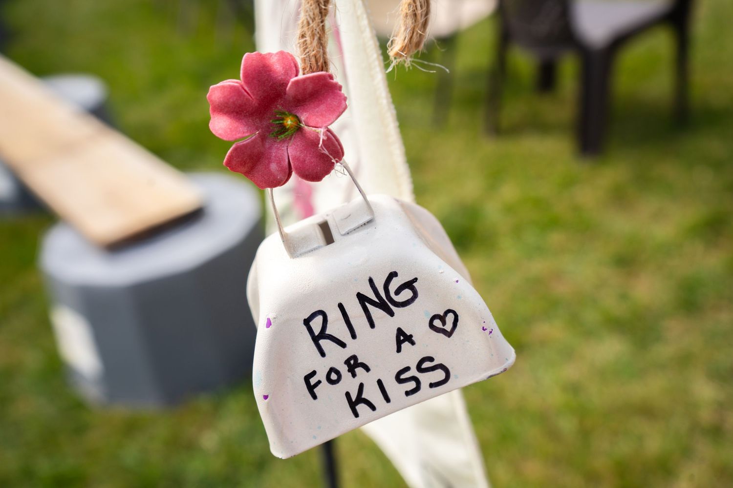 A decorative white cowbell with a pink flower reads Ring For A Kiss at an outdoor wedding ceremony.