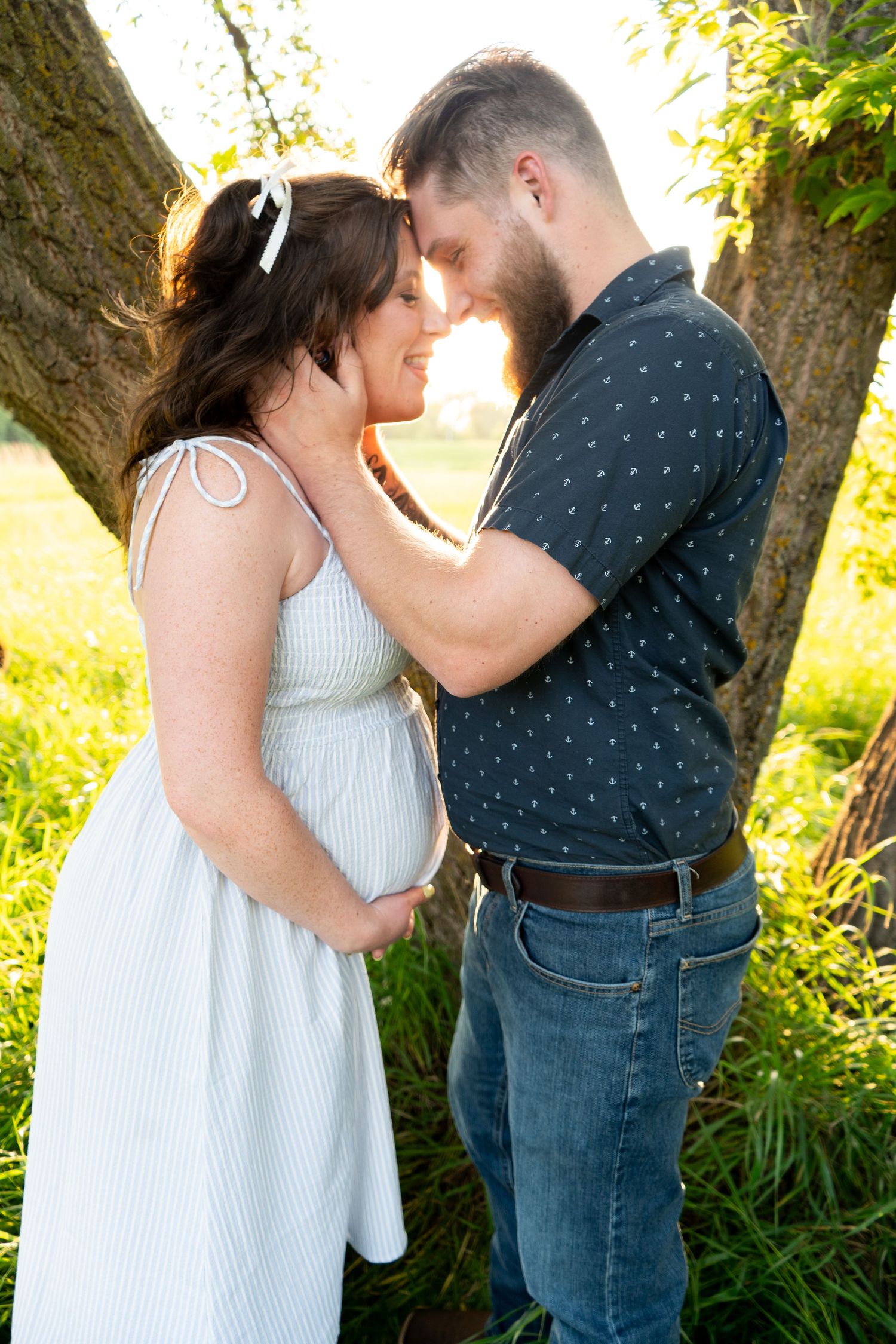 Pregnant couple sharing tender forehead touch near tree in golden sunlight during maternity session.