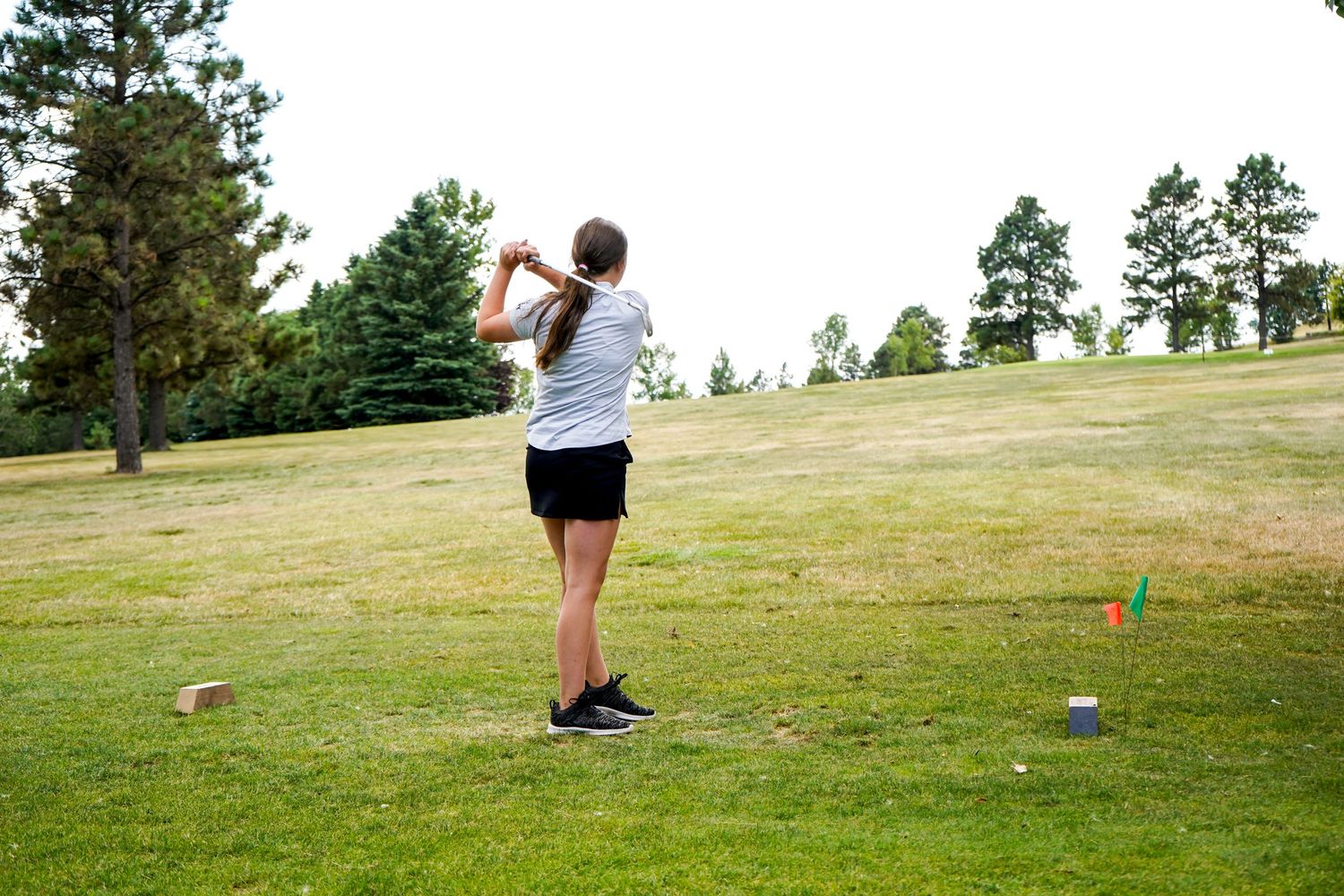 A golfer in a white shirt and black shorts completes a swing on a green golf course with pine trees.