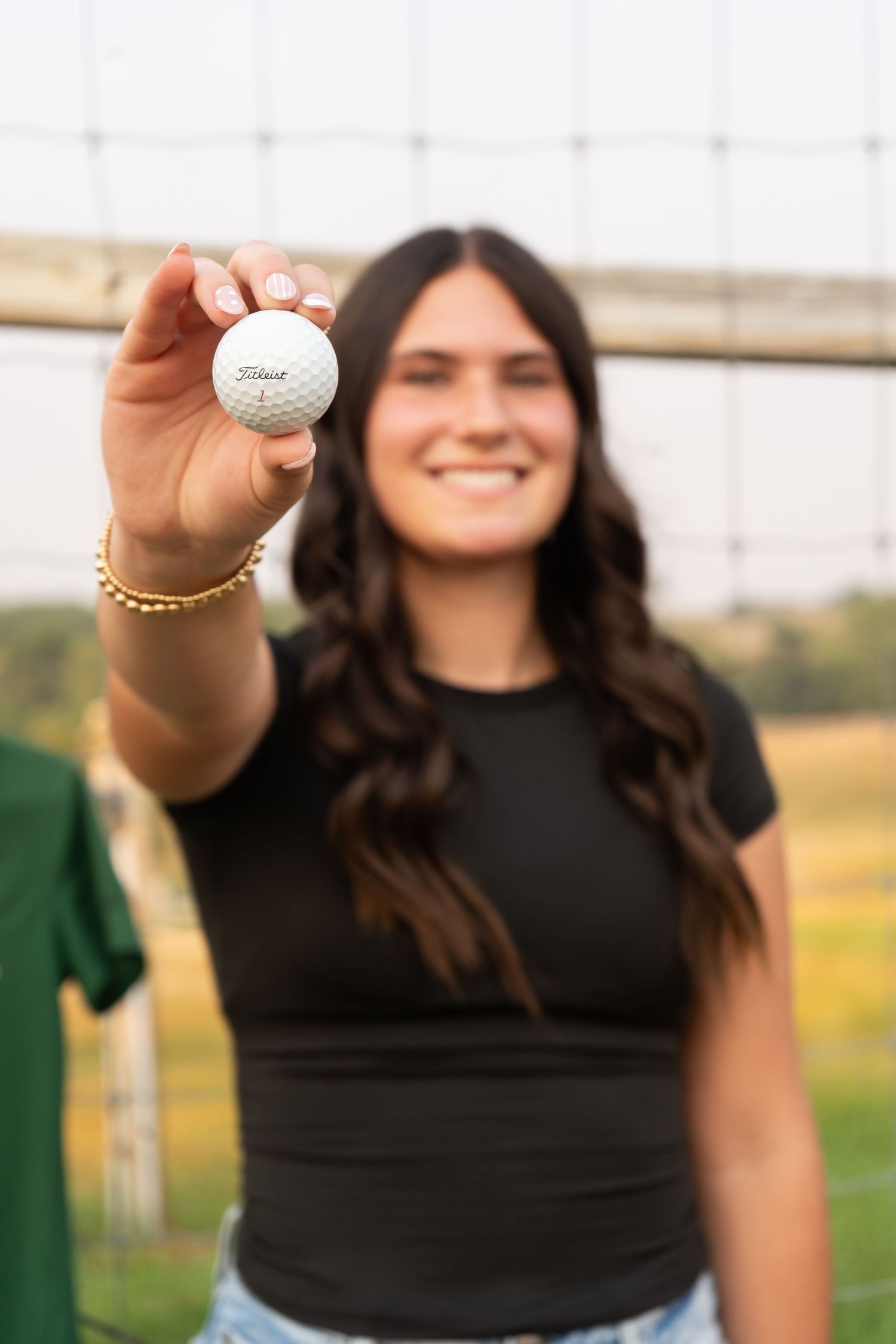 Smiling brunette girl holds up a Titleist golf ball toward the camera outdoors.