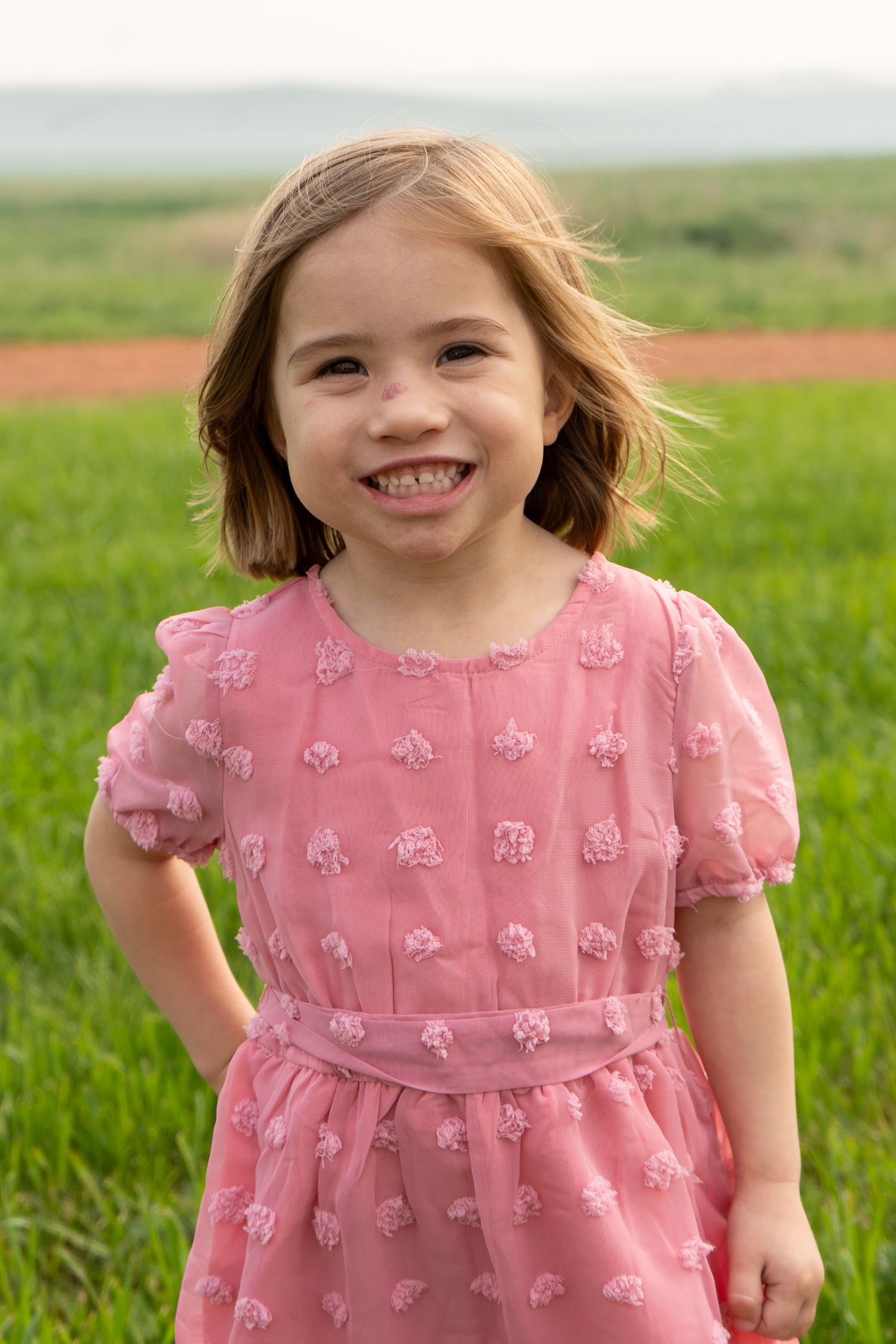 Smiling young girl in a pink textured dress standing in a green field.