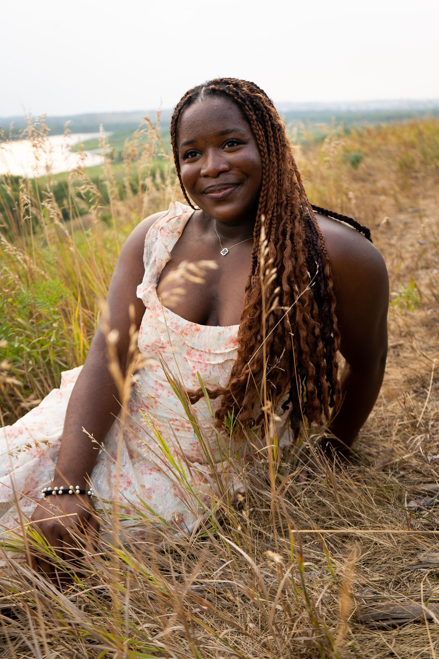 Happy young Black woman with long braids sitting in a golden grass field wearing a white floral sundress.