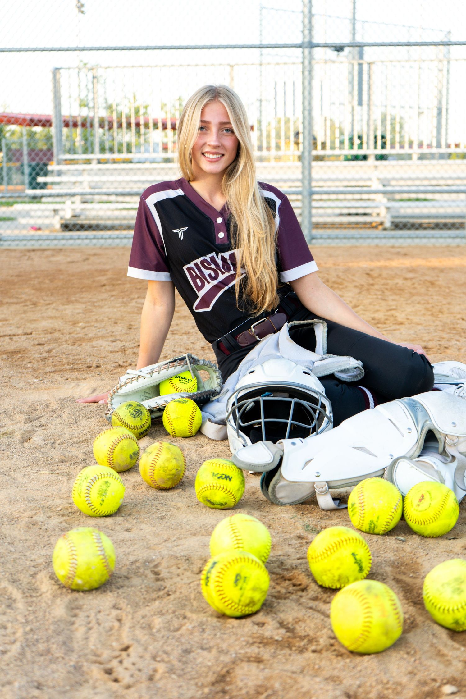 Blonde softball catcher posing with yellow softballs and catcher gear on a baseball diamond.