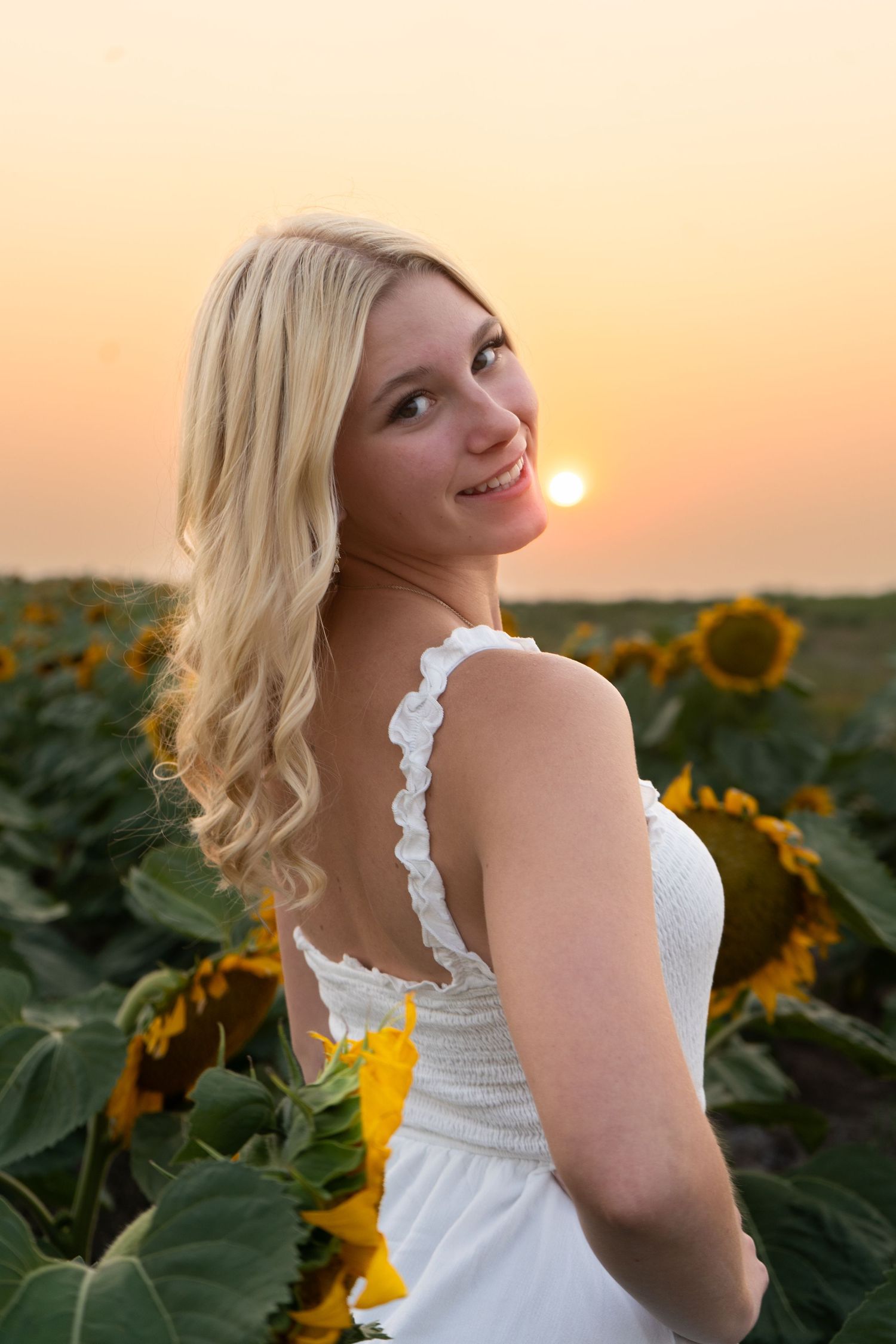 Blonde woman in white dress standing in sunflower field at golden sunset looking back.