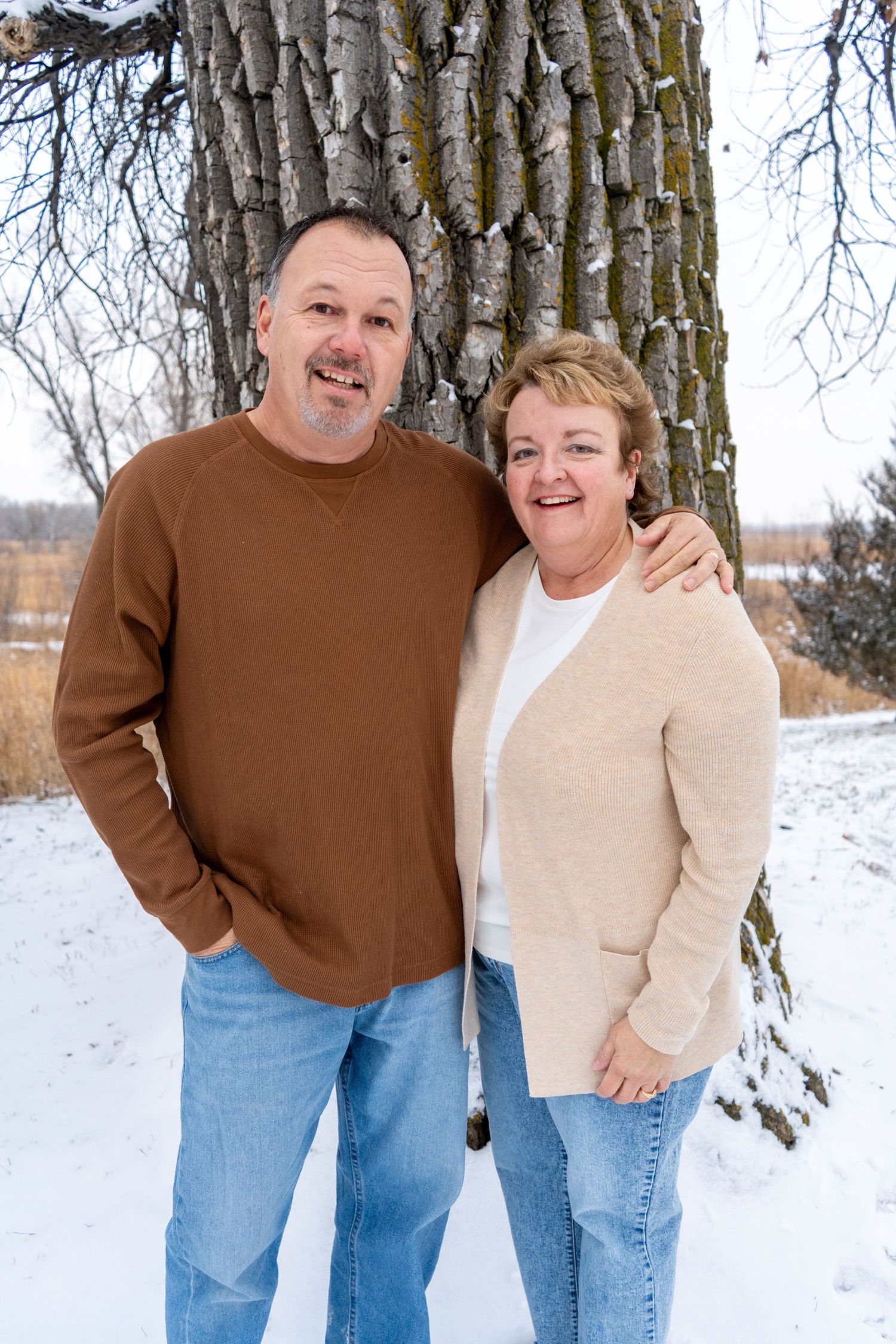 Happy couple posing together in snowy winter landscape near large tree, wearing casual jeans and sweaters.