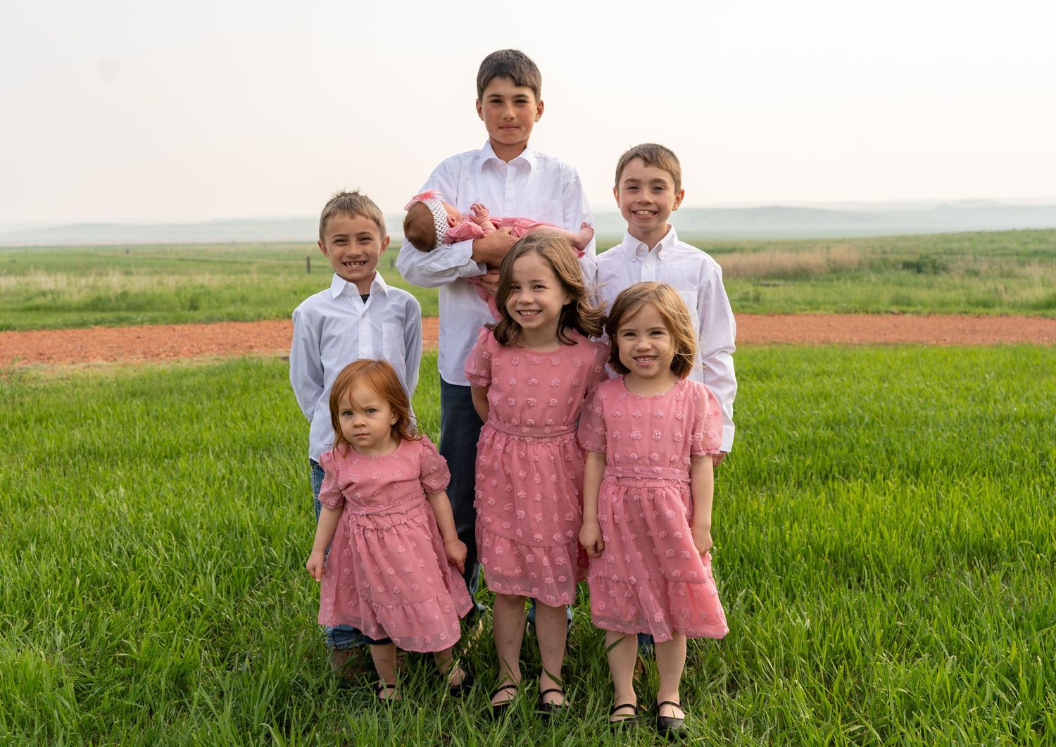 Six children in white and pink outfits pose together in a green grassy field.