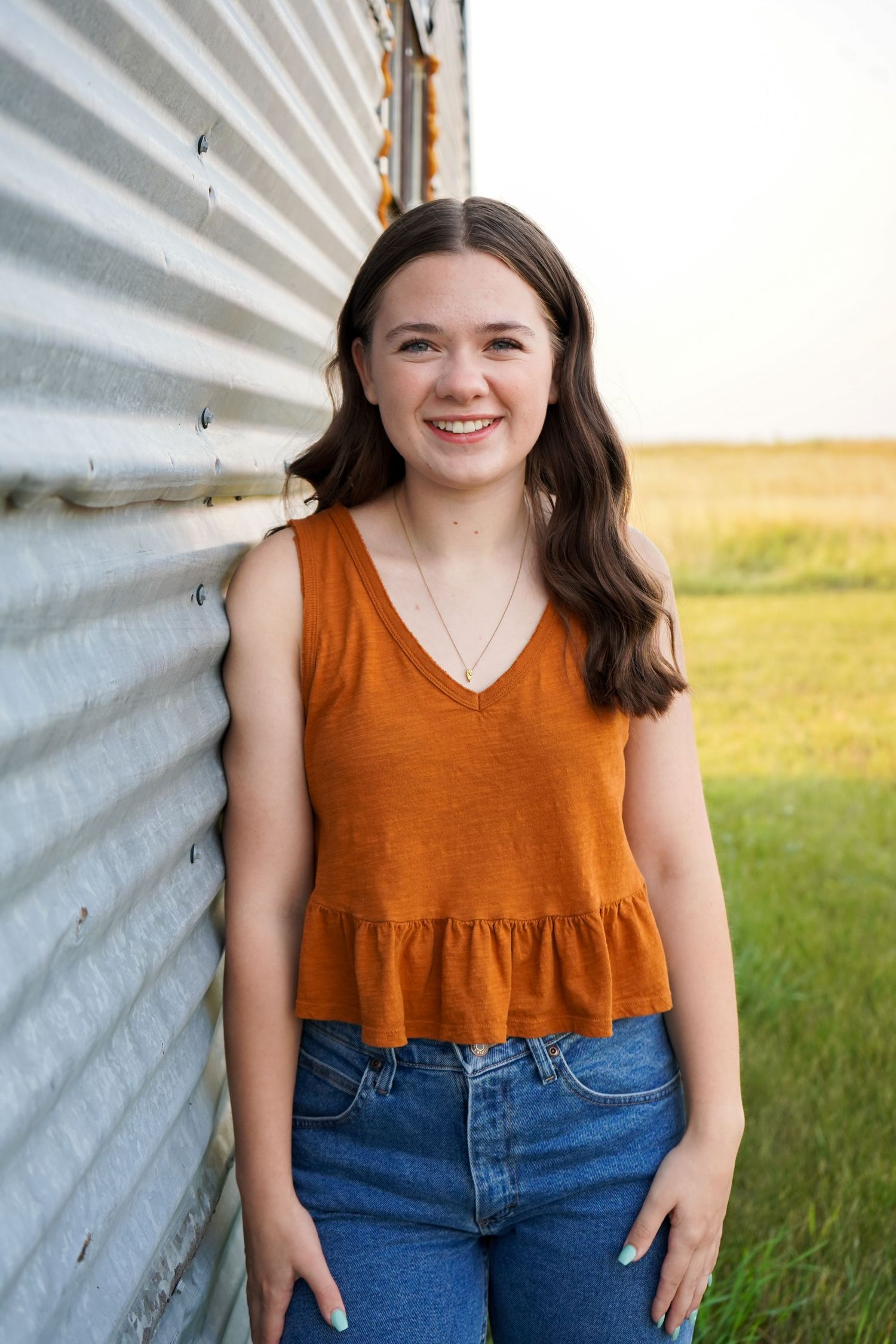 Smiling brunette teen girl in rust orange tank top leaning against corrugated metal wall.