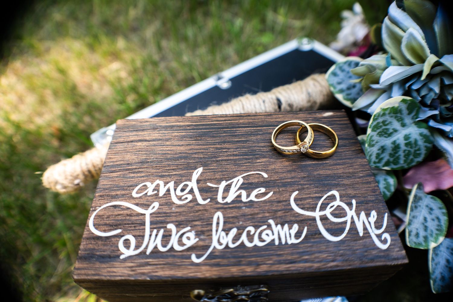 A rustic wooden box inscribed and the two become one displays two gold wedding rings beside a floral bouquet.