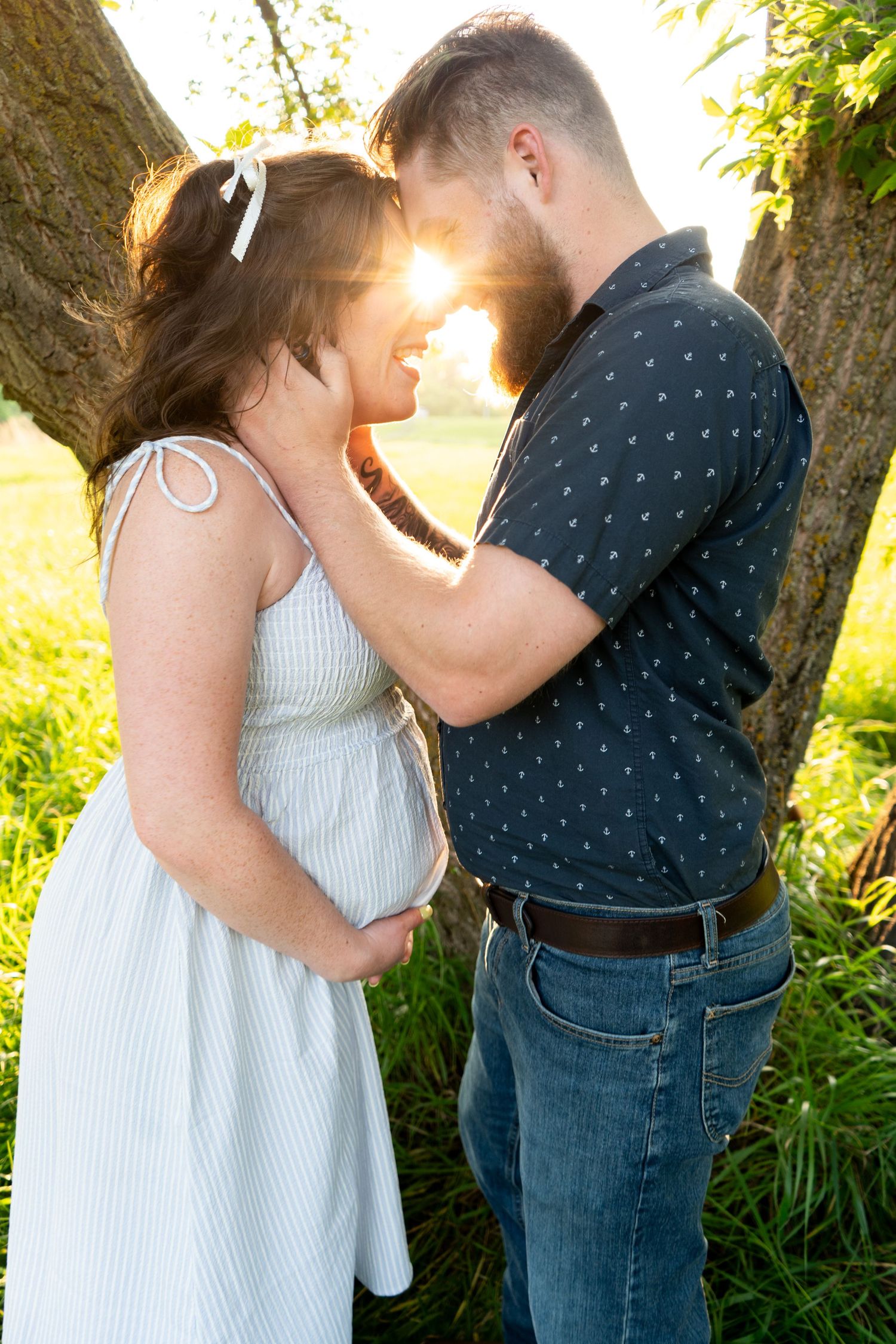 Expectant couple embracing near tree with sunlight shining between them during outdoor maternity shoot.
