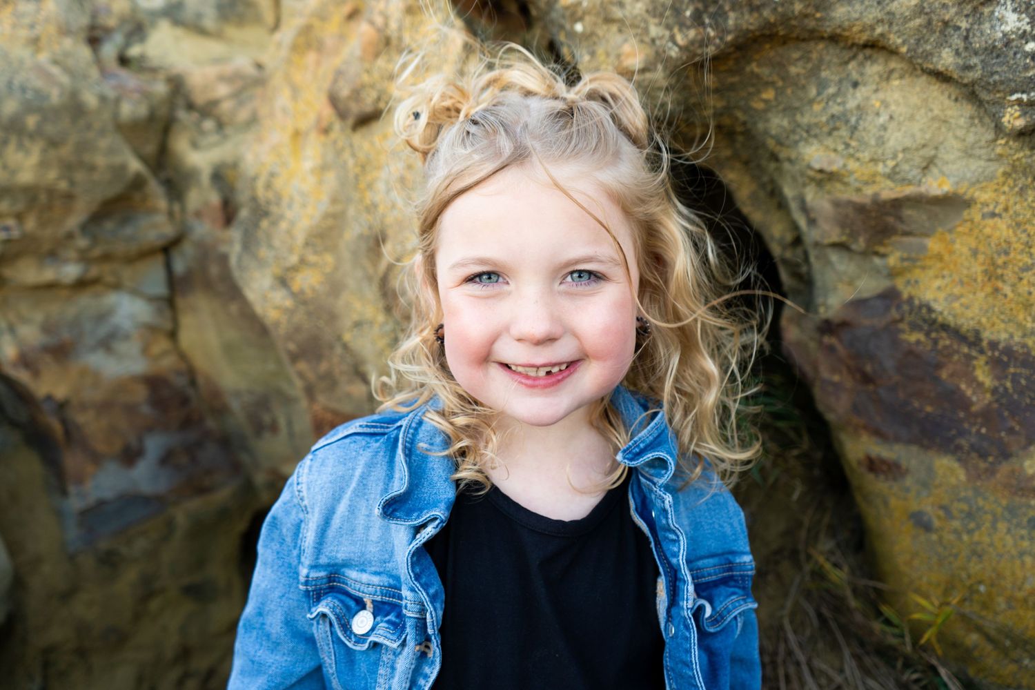 Cute blonde girl with curly hair smiling in denim jacket and black shirt near rocky backdrop.