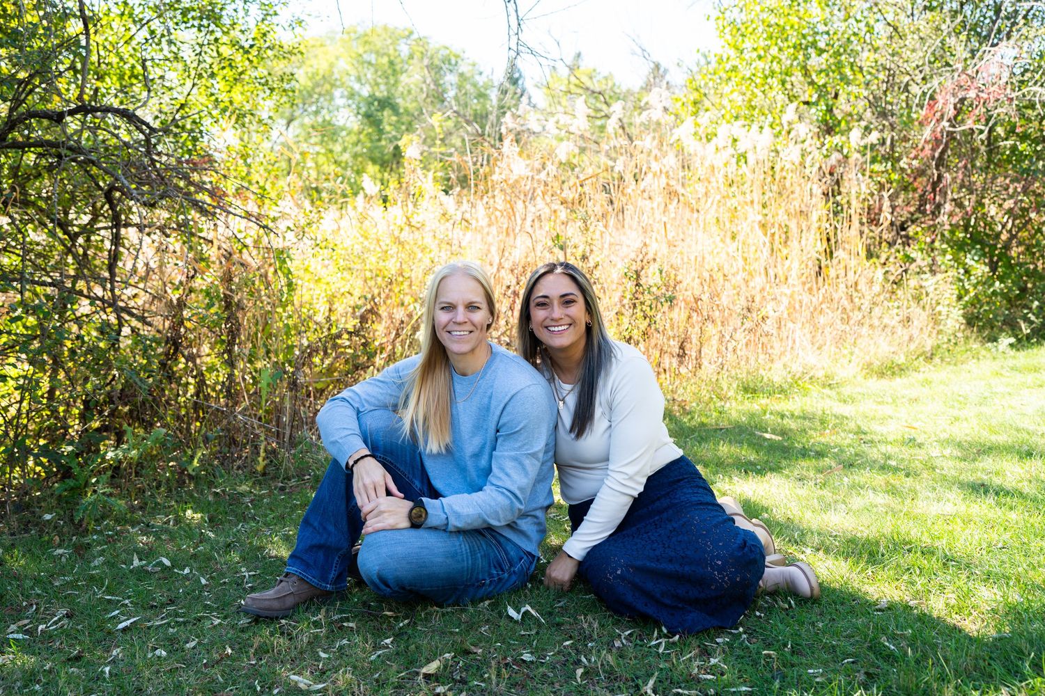 Two women sit together smiling on grass surrounded by fall foliage.