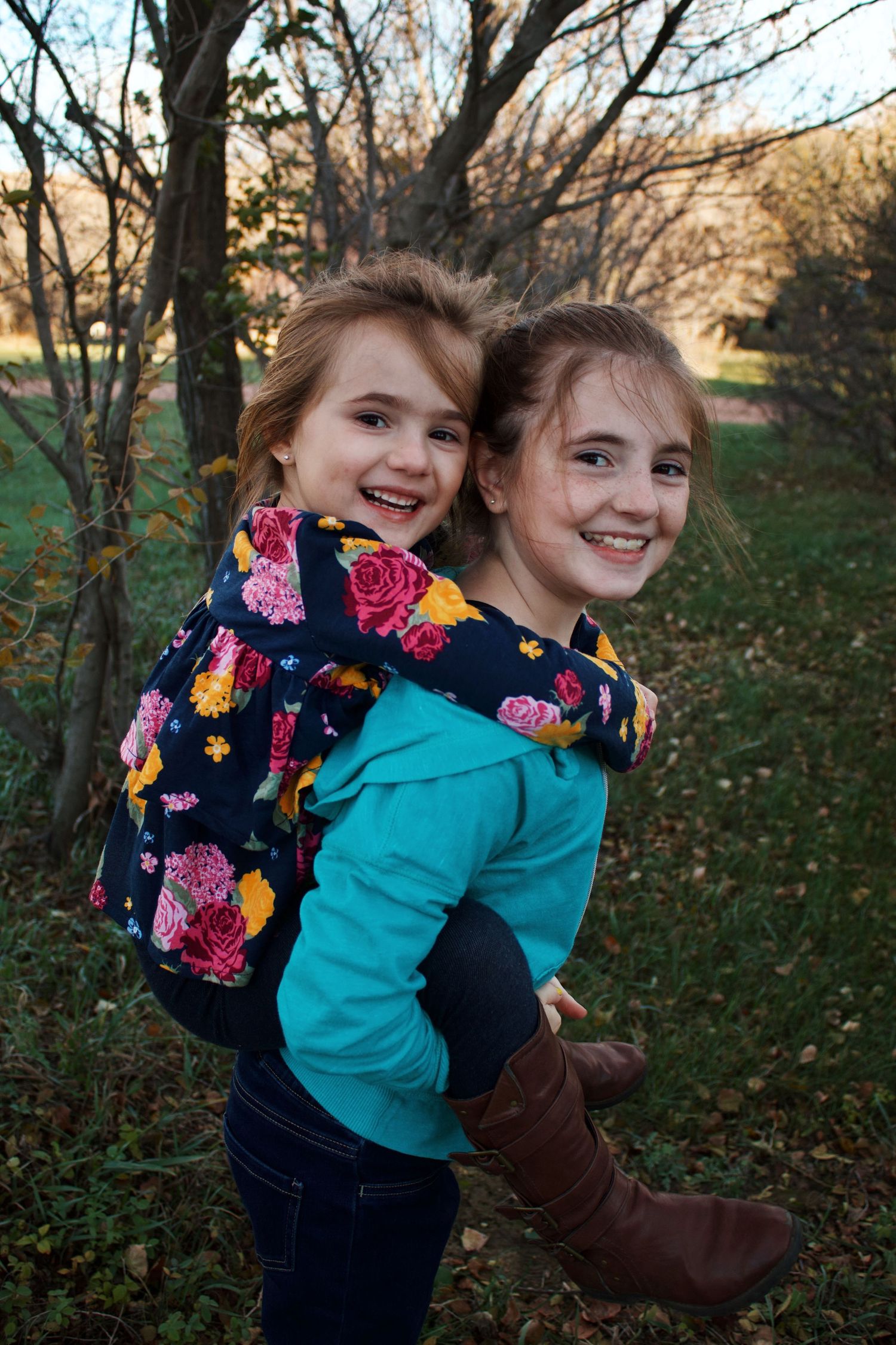 Two young girls laughing as older sister gives younger a piggyback ride outdoors.