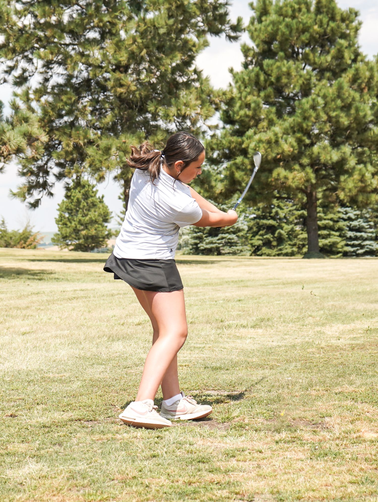 A female golfer in a white shirt and black skort follows through on a golf swing on a sunny course.