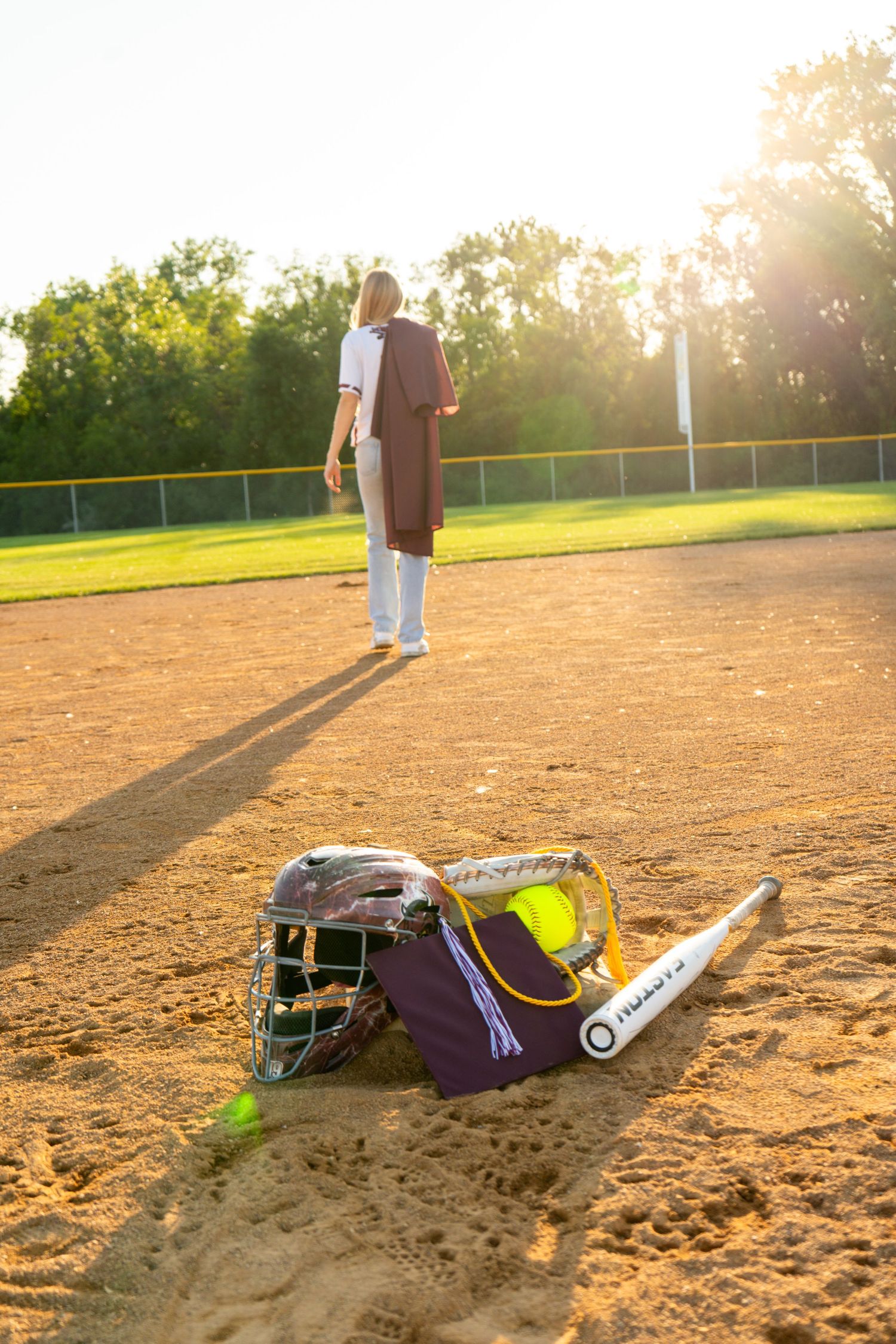 Softball player walking away on a sunlit diamond with catcher gear and bat bag in the foreground.