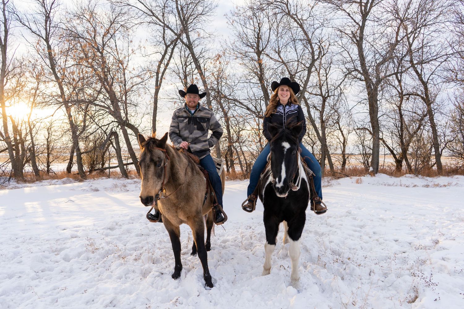 Couple on horseback riding through a snowy landscape at sunset wearing black cowboy hats.