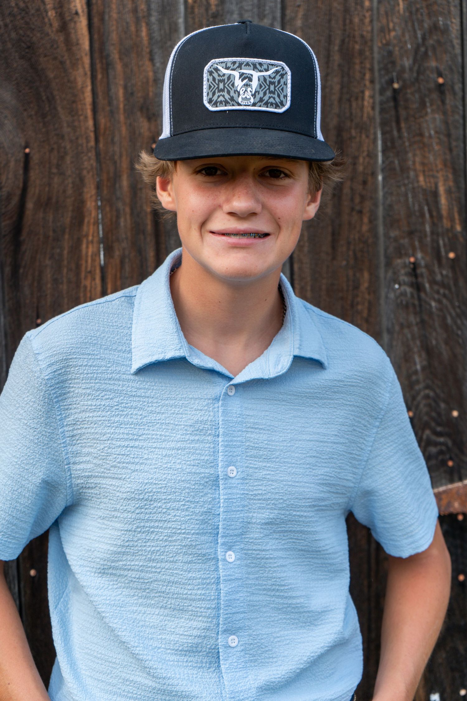 Smiling teenage boy wearing a light blue button-up shirt and black snapback cap in front of a wooden barn wall.