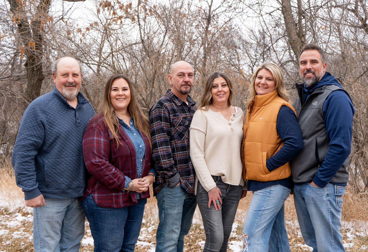 Group of six adults smiling together outdoors in winter with bare trees in background.