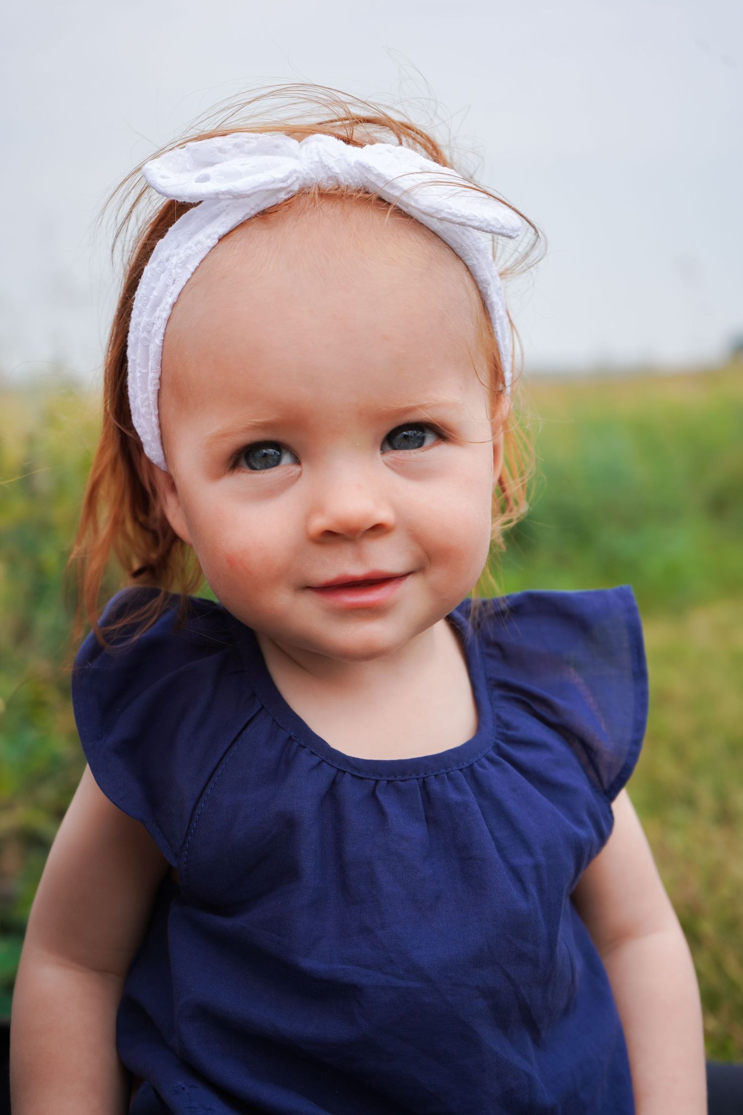 Cute red-haired baby girl wearing a white bow headband and navy blue dress smiling outdoors.