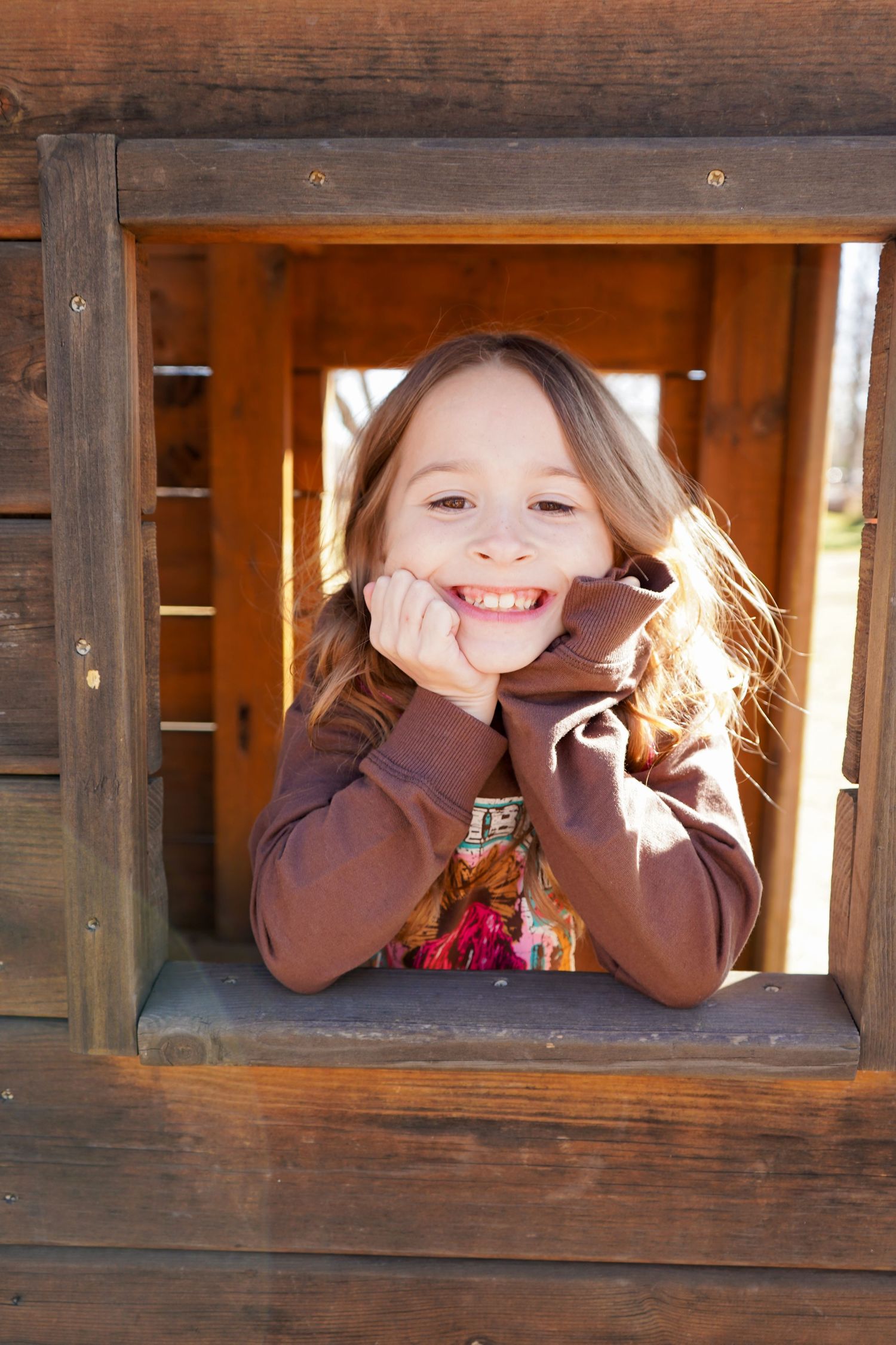 Happy young girl with long hair leaning through wooden playhouse window smiling.