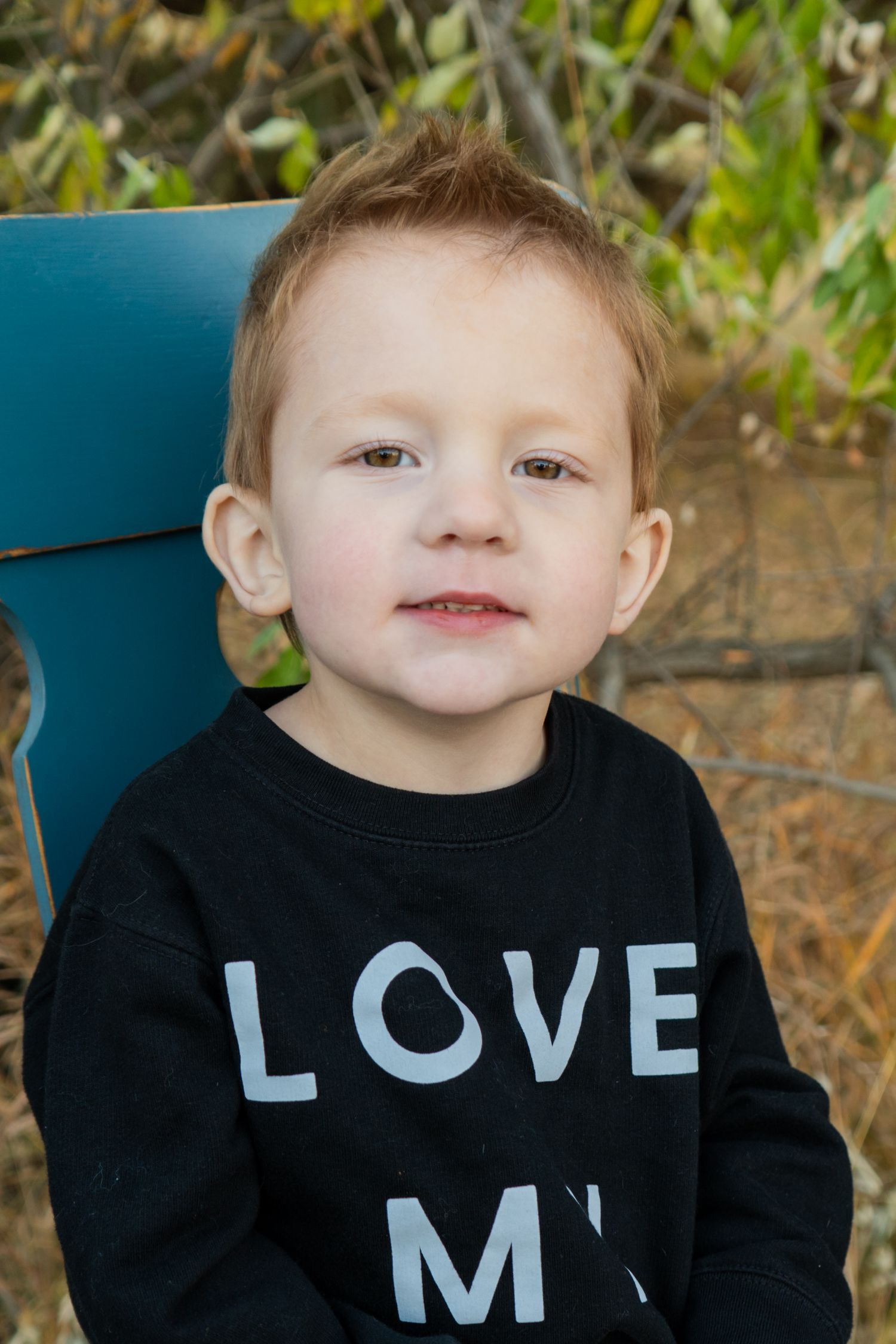 Adorable red-haired toddler boy wearing a black Love sweatshirt sitting in a blue chair outdoors.