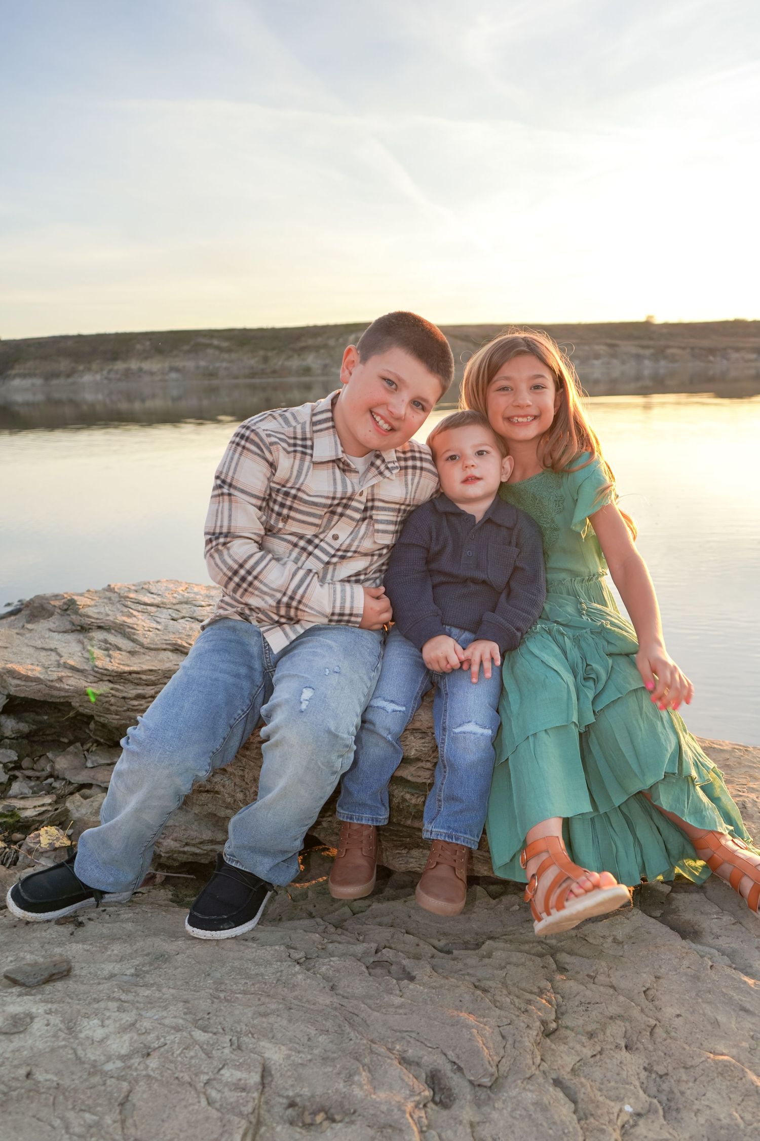 Three siblings sit together on rocky lakeside ledge at golden hour, smiling for a portrait.