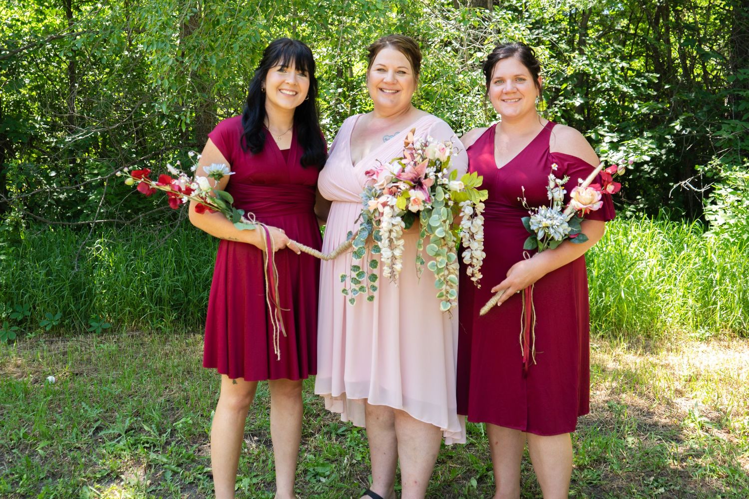 A bride in pink stands between two bridesmaids in burgundy dresses holding bouquets in a garden setting.