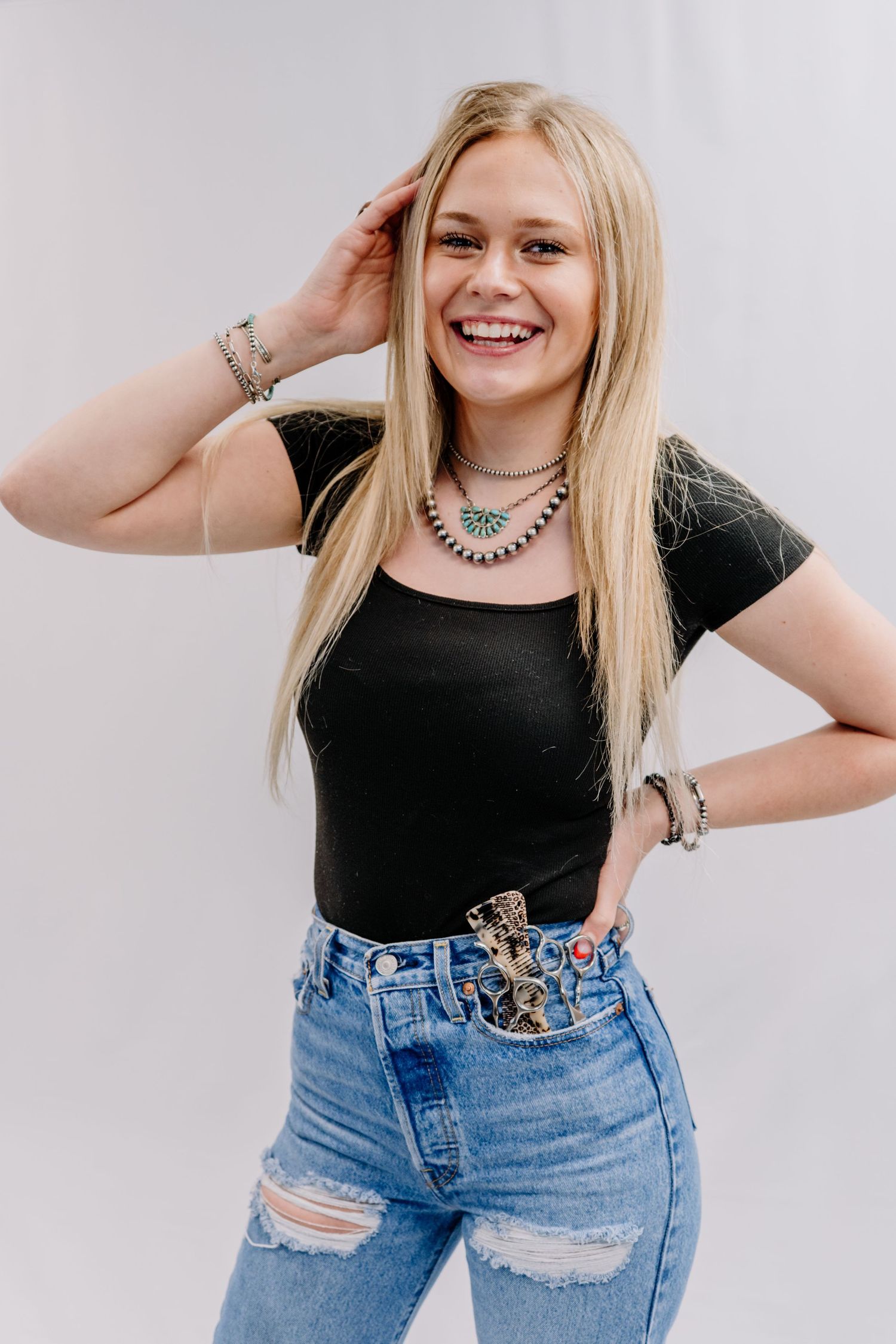 Smiling blonde woman in black crop top, ripped jeans, and turquoise necklace posing against white background.