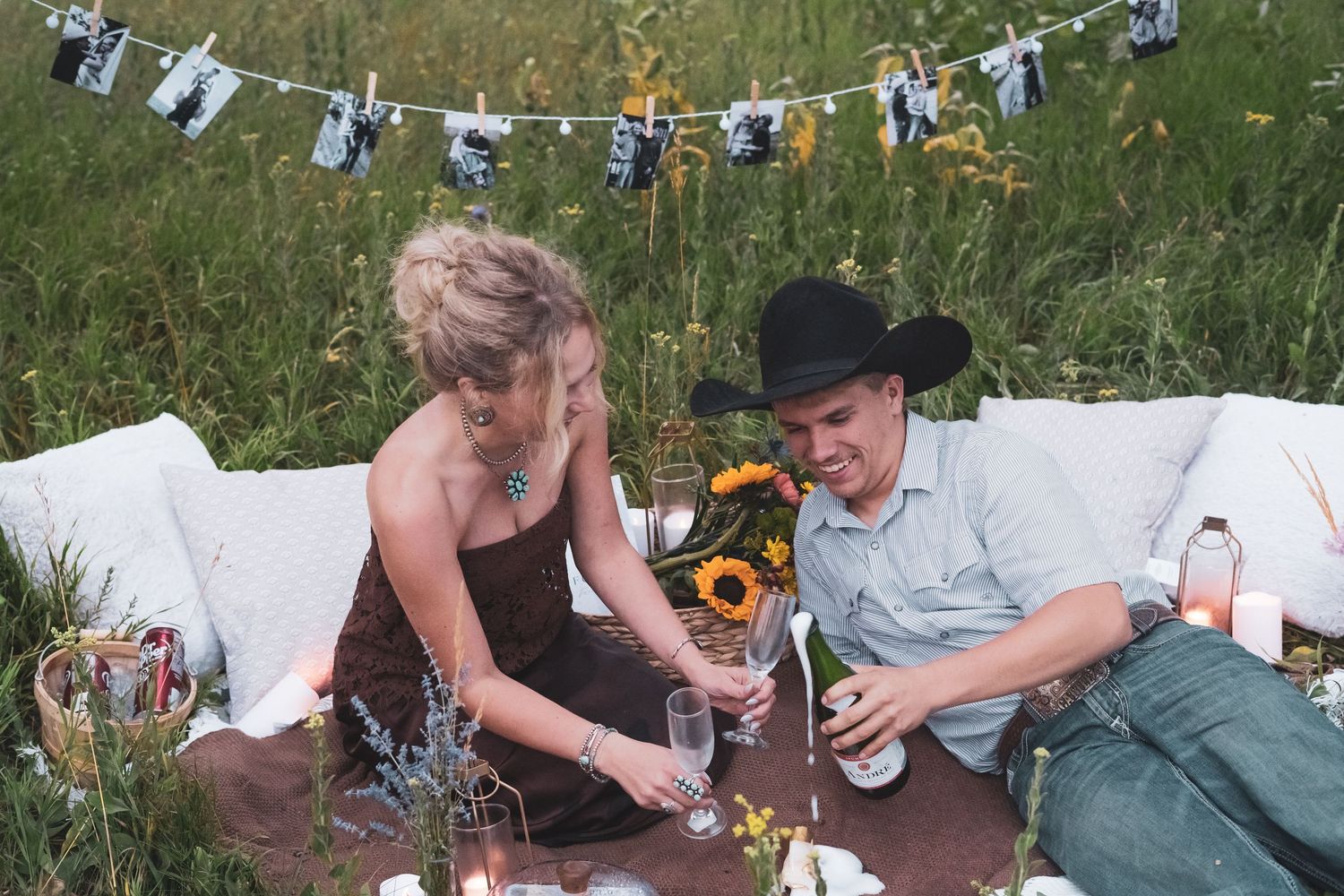 Couple sharing a romantic picnic in a wildflower field with sunflowers and a photo garland backdrop.