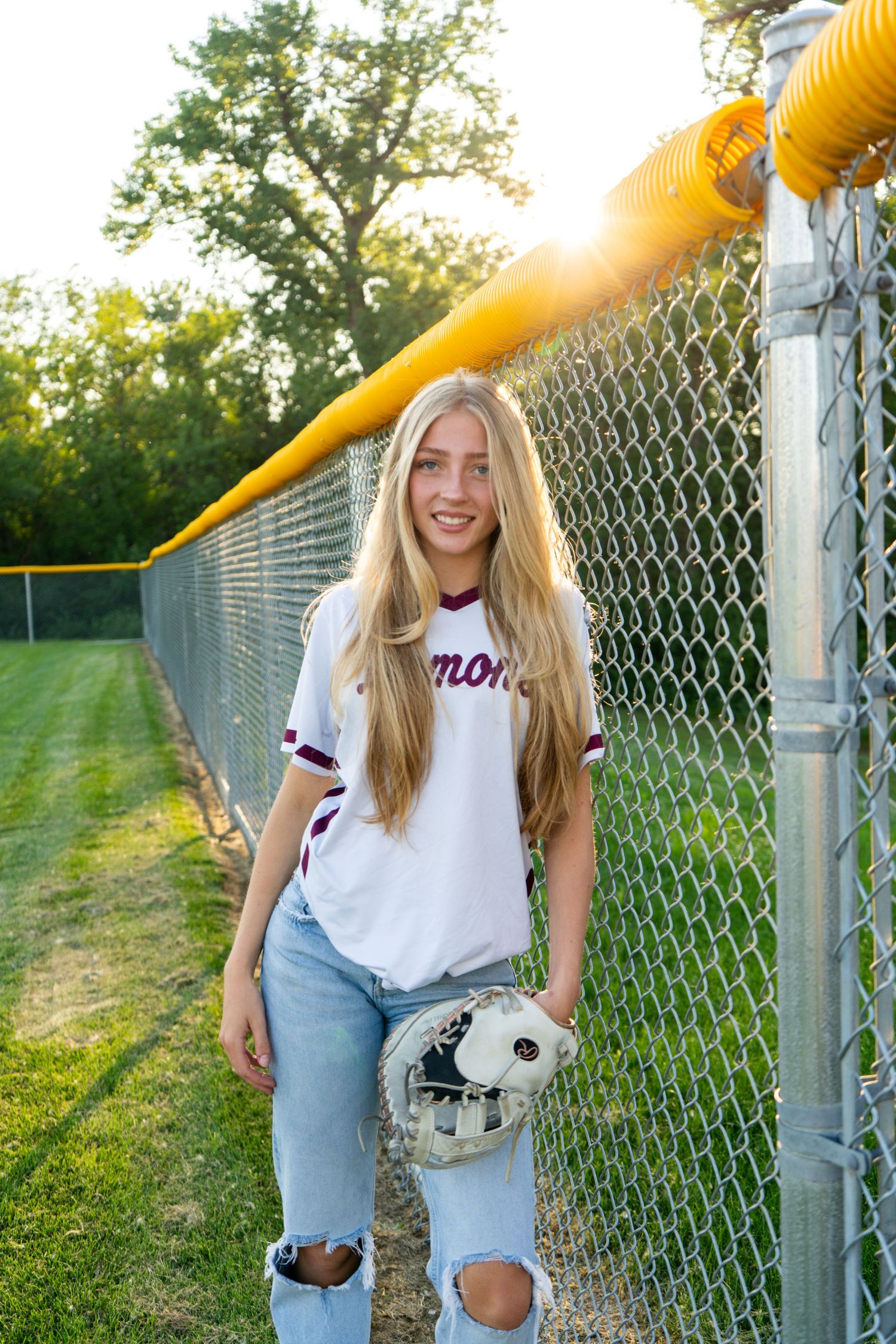 Smiling blonde softball player in white jersey and ripped jeans holding a glove by a chain-link fence.