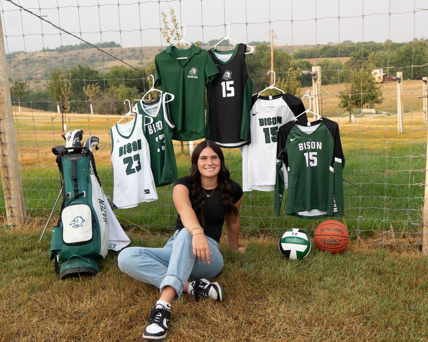 Senior girl sits by sports jerseys and equipment on a fence in a rural setting.