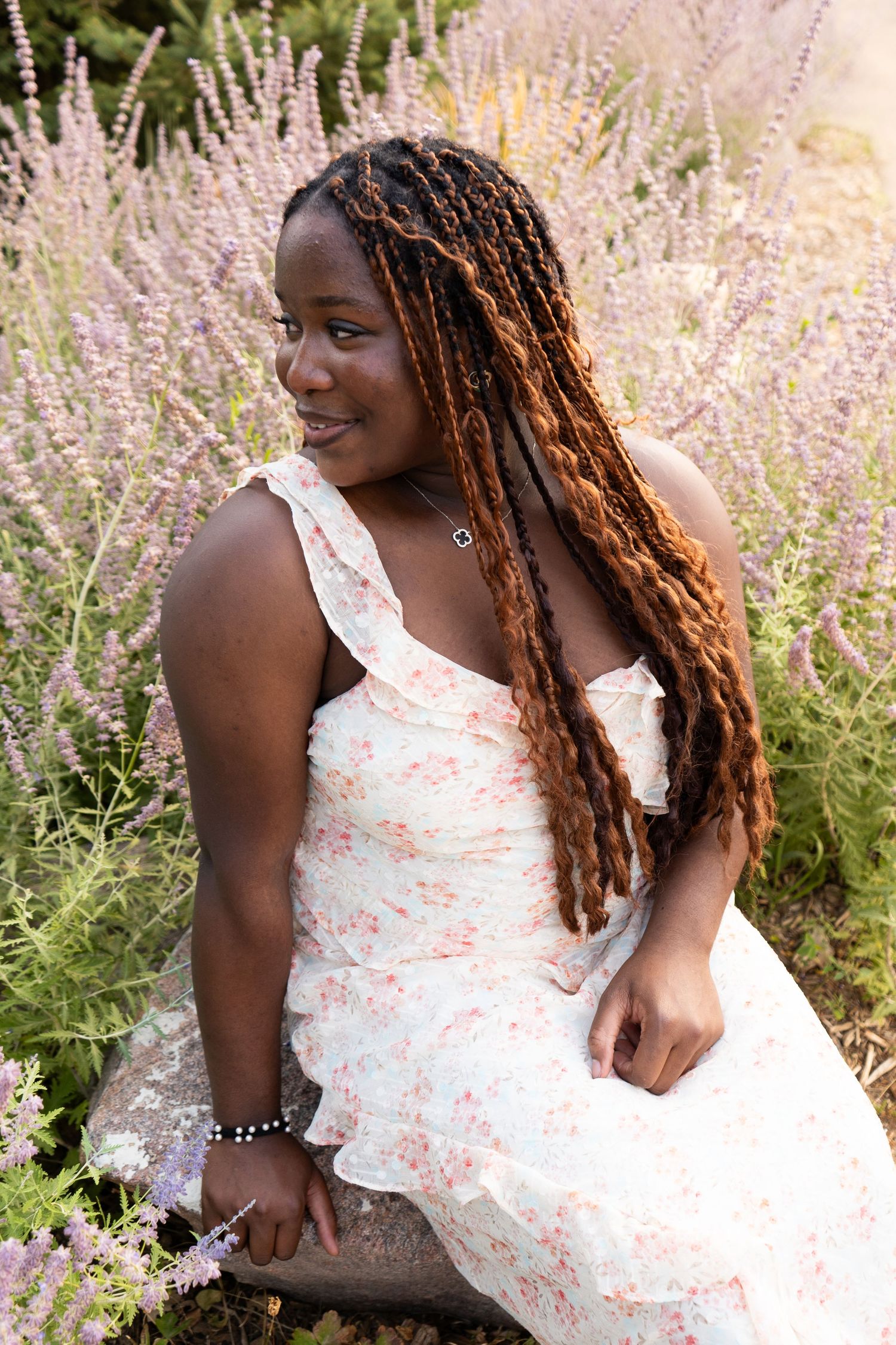Young Black woman with long auburn box braids sitting among purple lavender flowers in a floral dress.