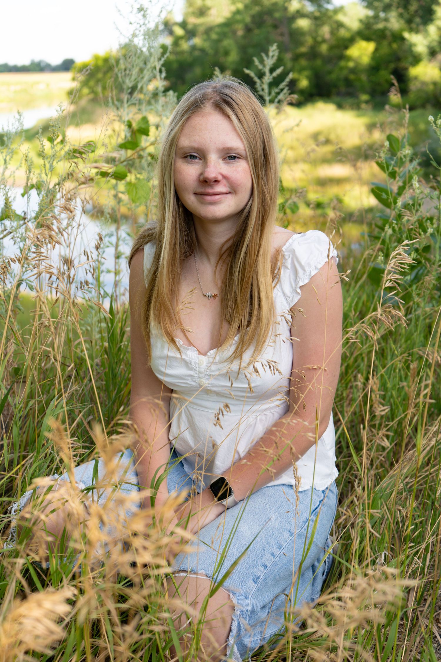 Smiling blonde teen girl sits in tall grass near a peaceful pond outdoors.