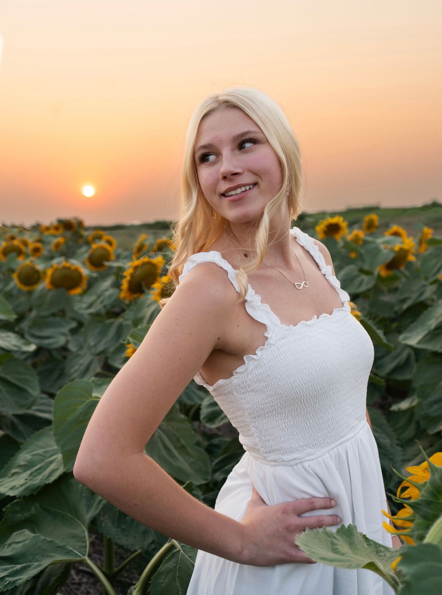 Smiling blonde woman in white dress posing in sunflower field at golden hour sunset.