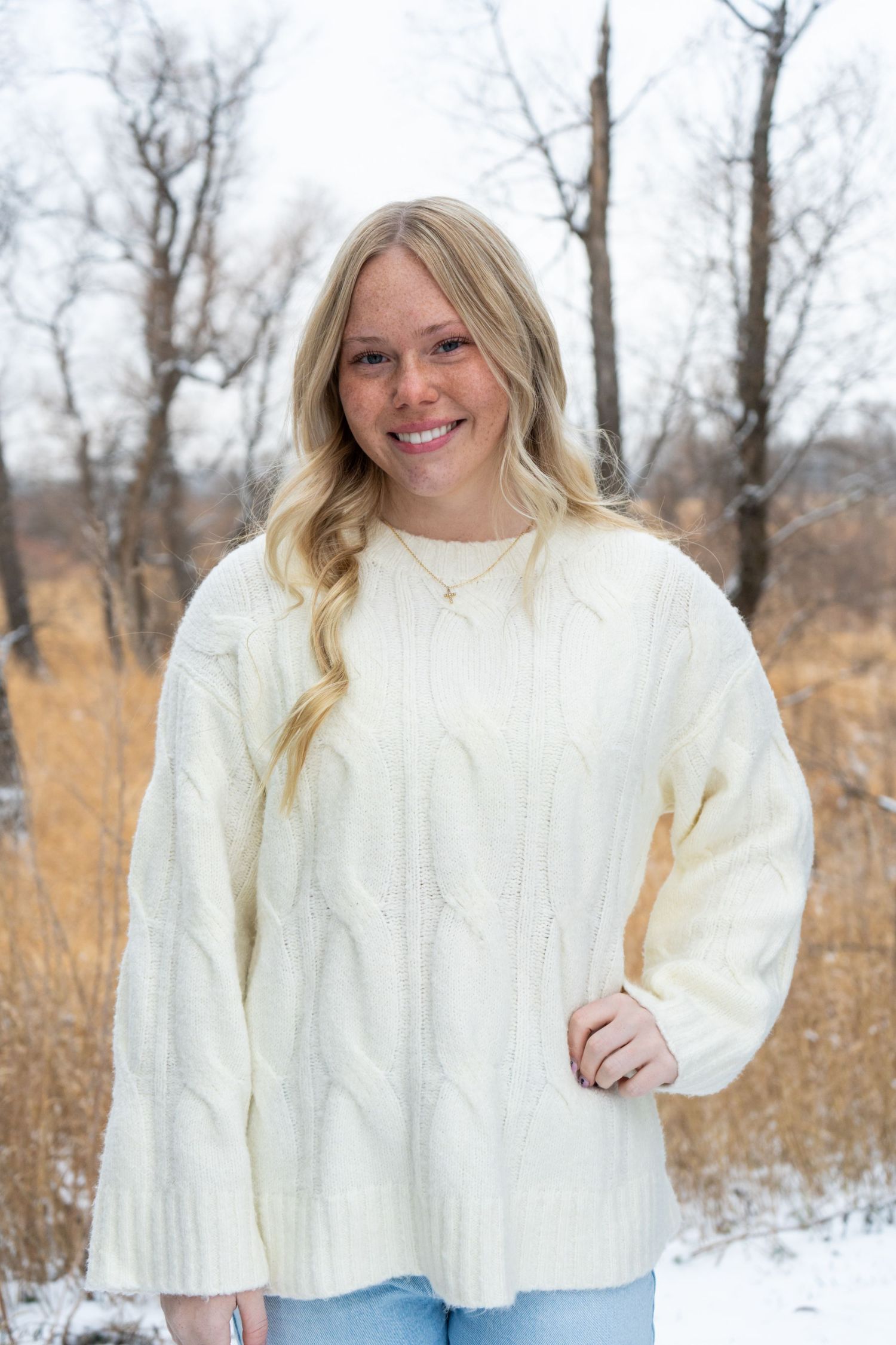 Young blonde woman smiling outdoors in cream cable-knit sweater with snowy winter background.
