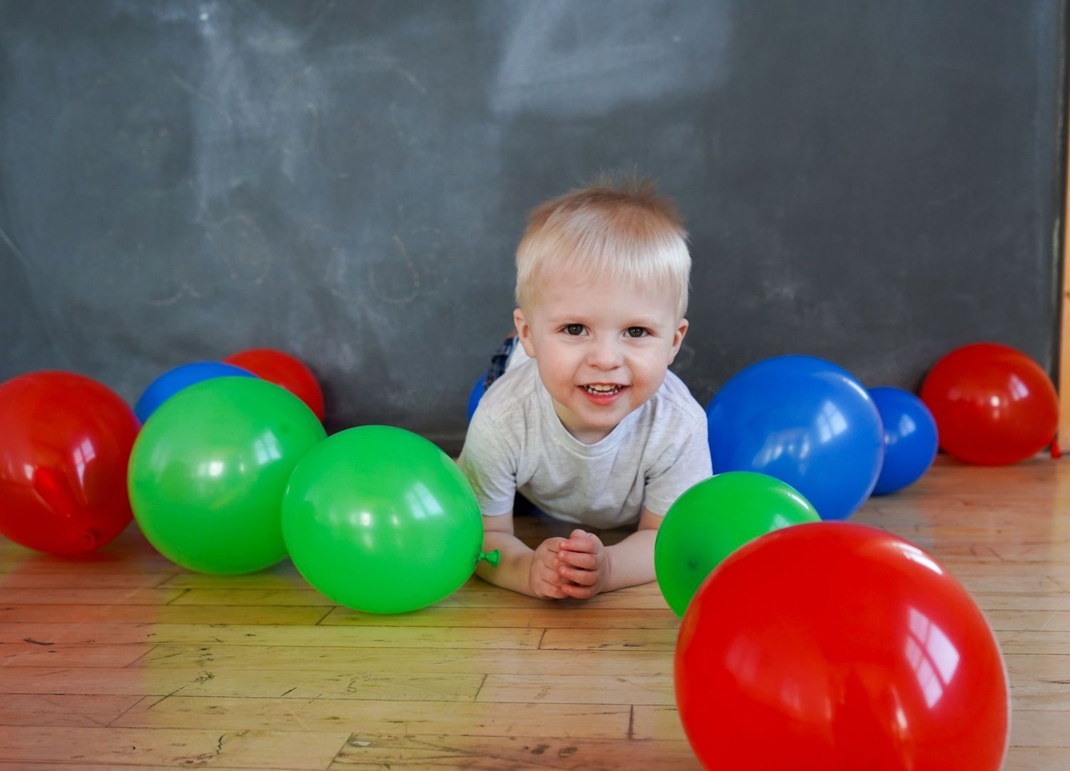 Smiling baby boy lying among colorful red, green, and blue balloons on a hardwood floor.
