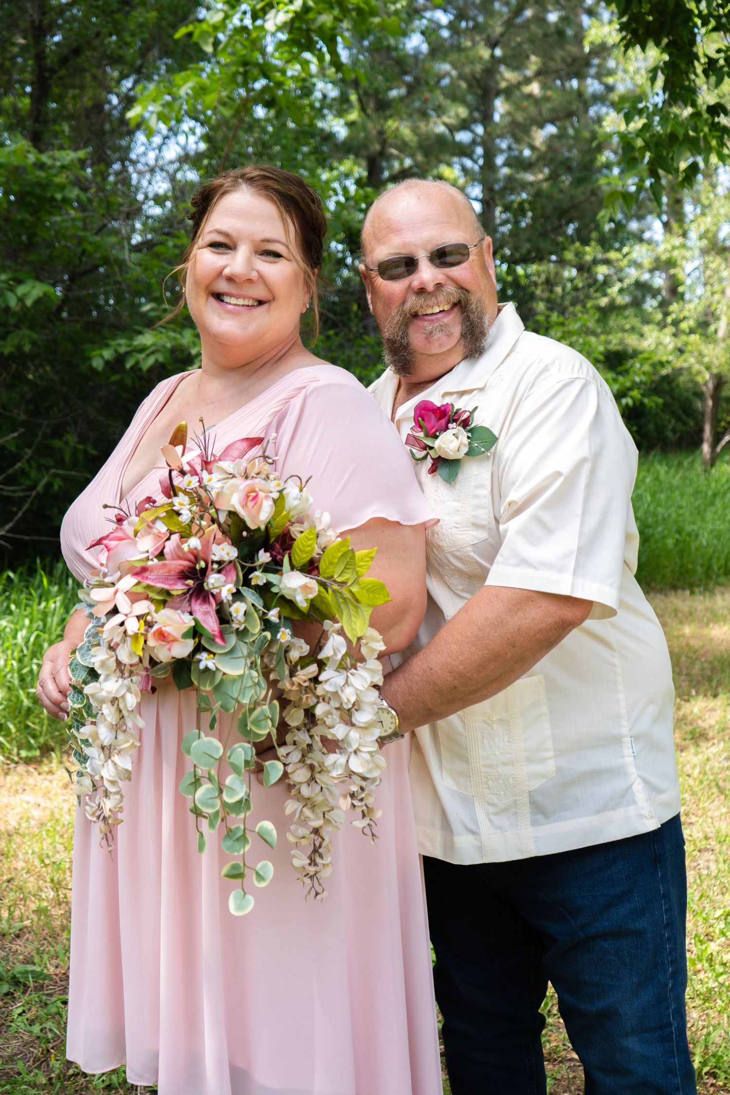 A smiling bride in a pink dress holds a cascading floral bouquet beside her groom in a white shirt outdoors.