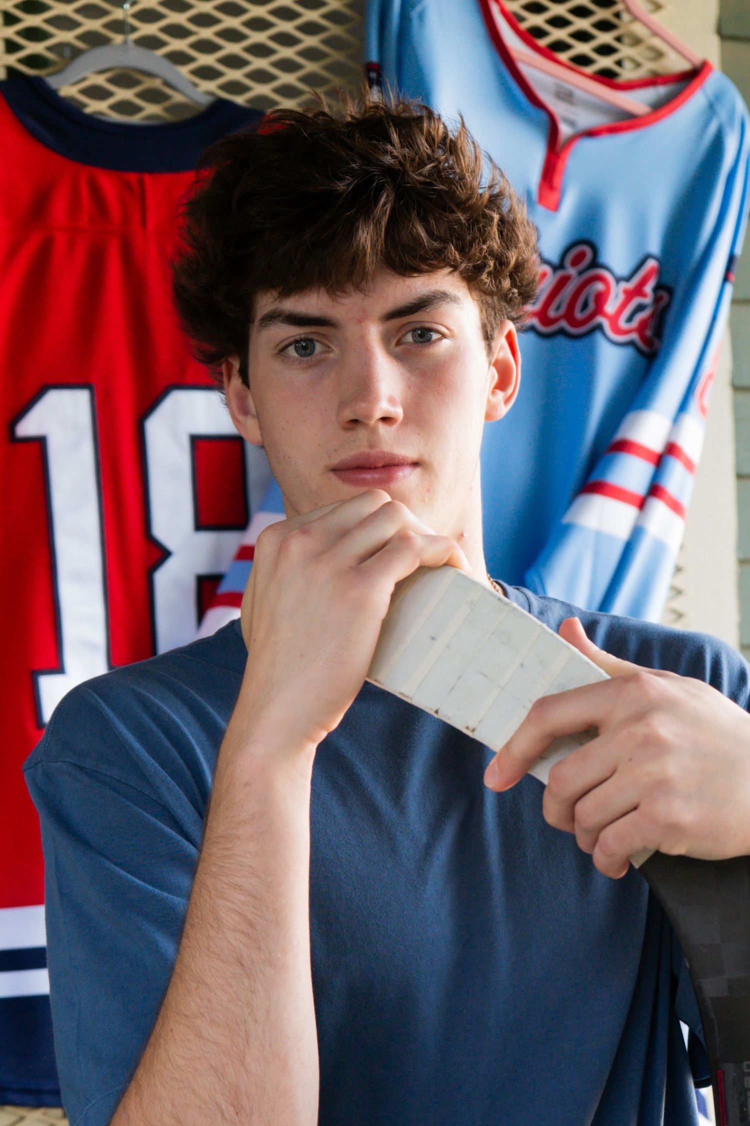 Teen boy holding hockey stick in front of sports jerseys in locker room setting.