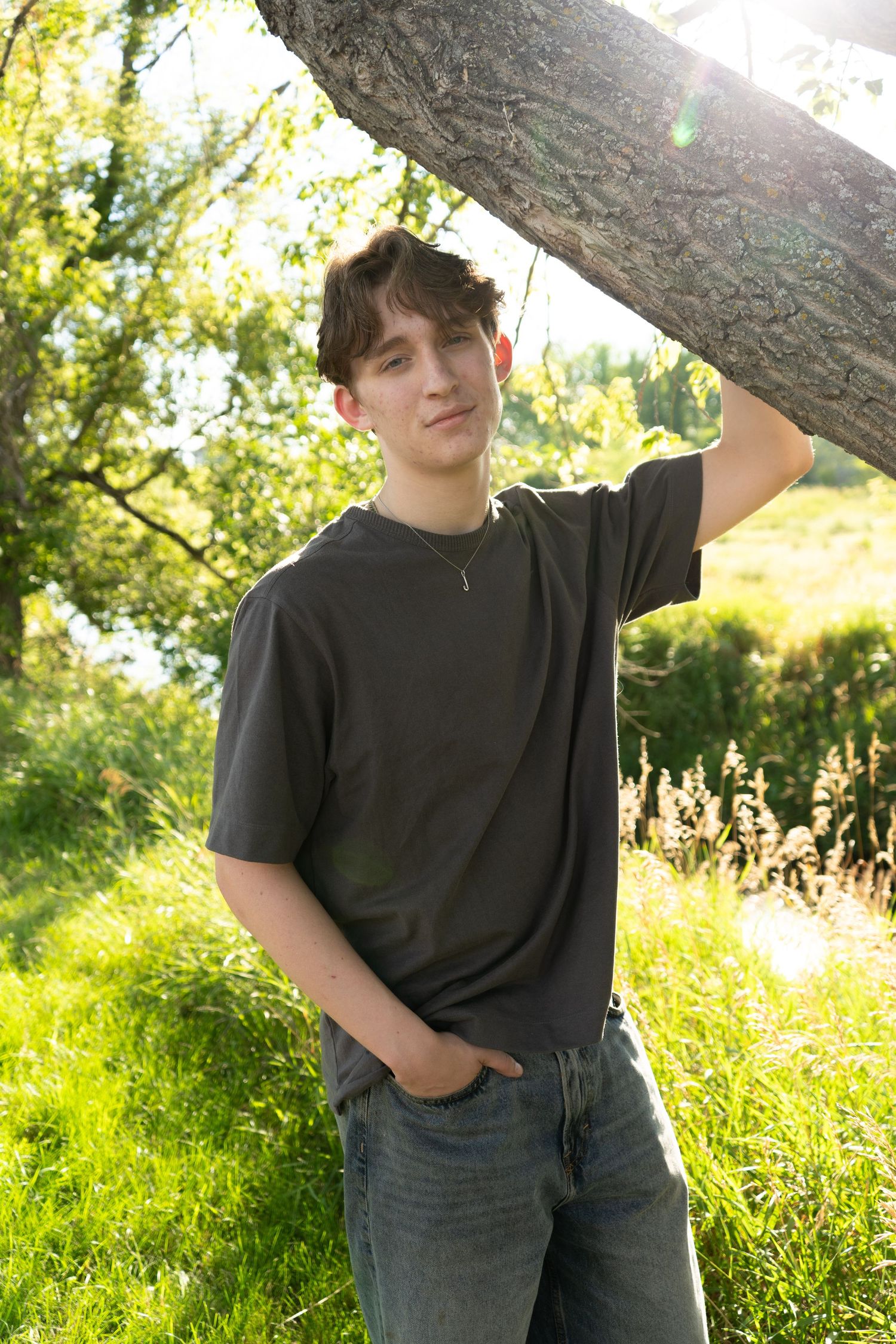 Teen boy in gray t-shirt leaning against tree trunk in sunny green outdoor setting.