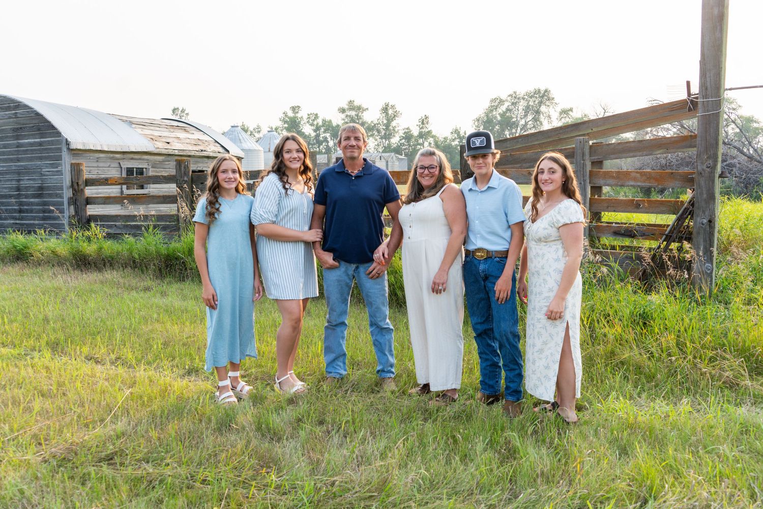 A family of six poses together in a sunny green field near a rustic wooden fence.