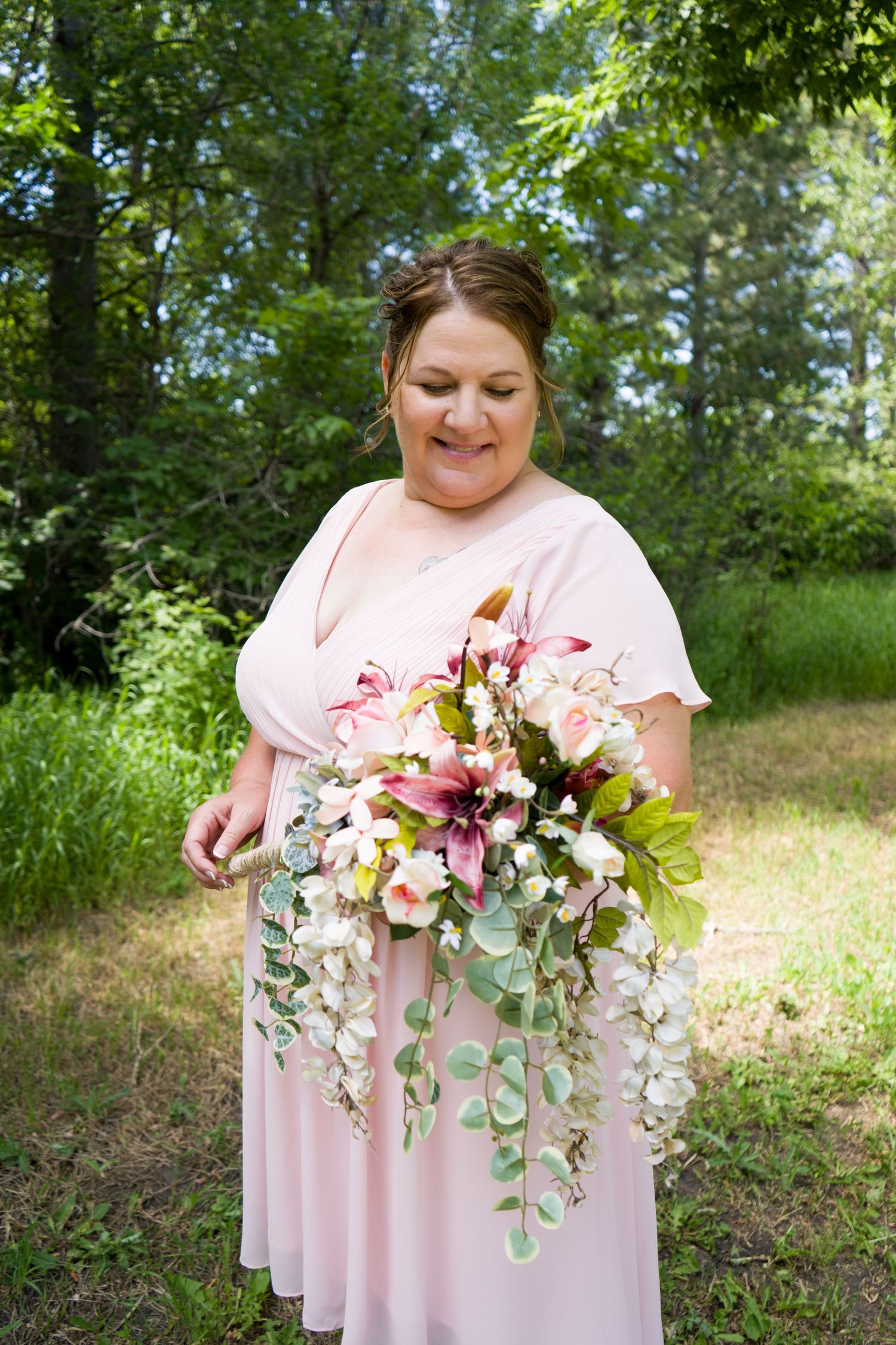 Smiling bride in pink dress admiring cascading floral bouquet with lilies and orchids in outdoor garden setting.