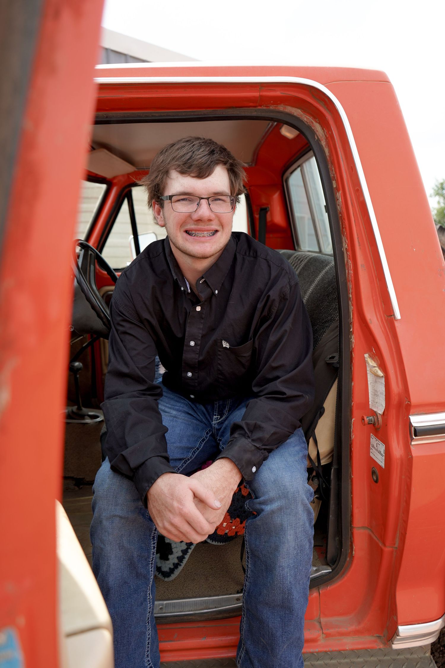 Smiling young man with braces sitting in doorway of vintage red pickup truck.