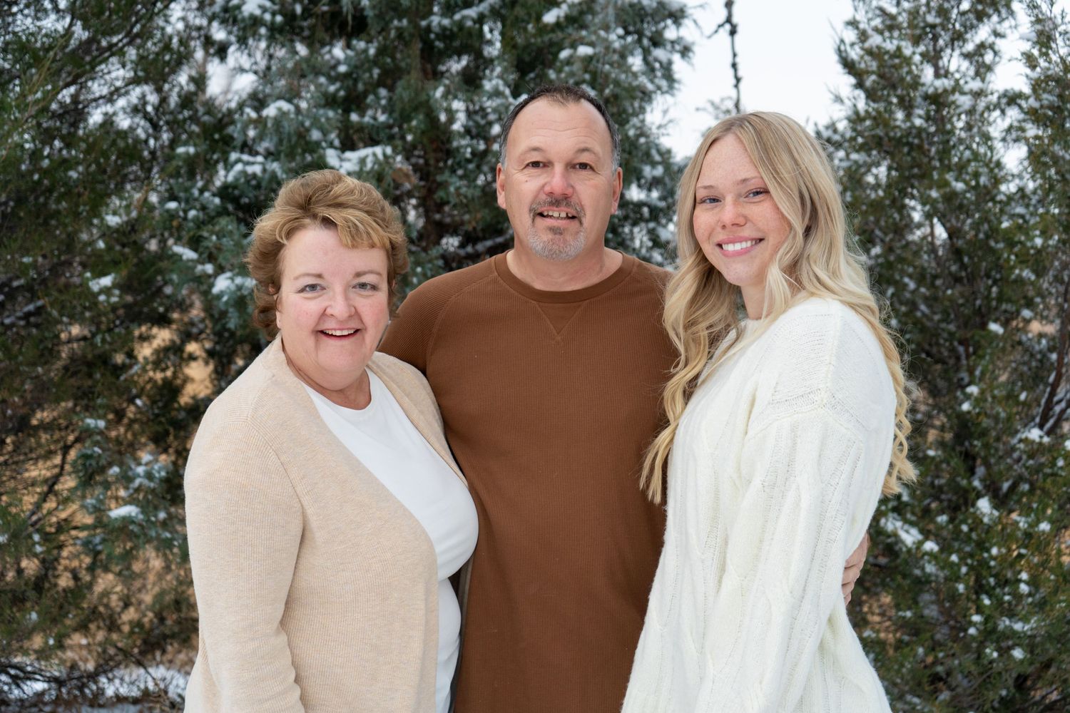 Family of three smiling together outdoors in winter with snow-covered evergreen trees behind.