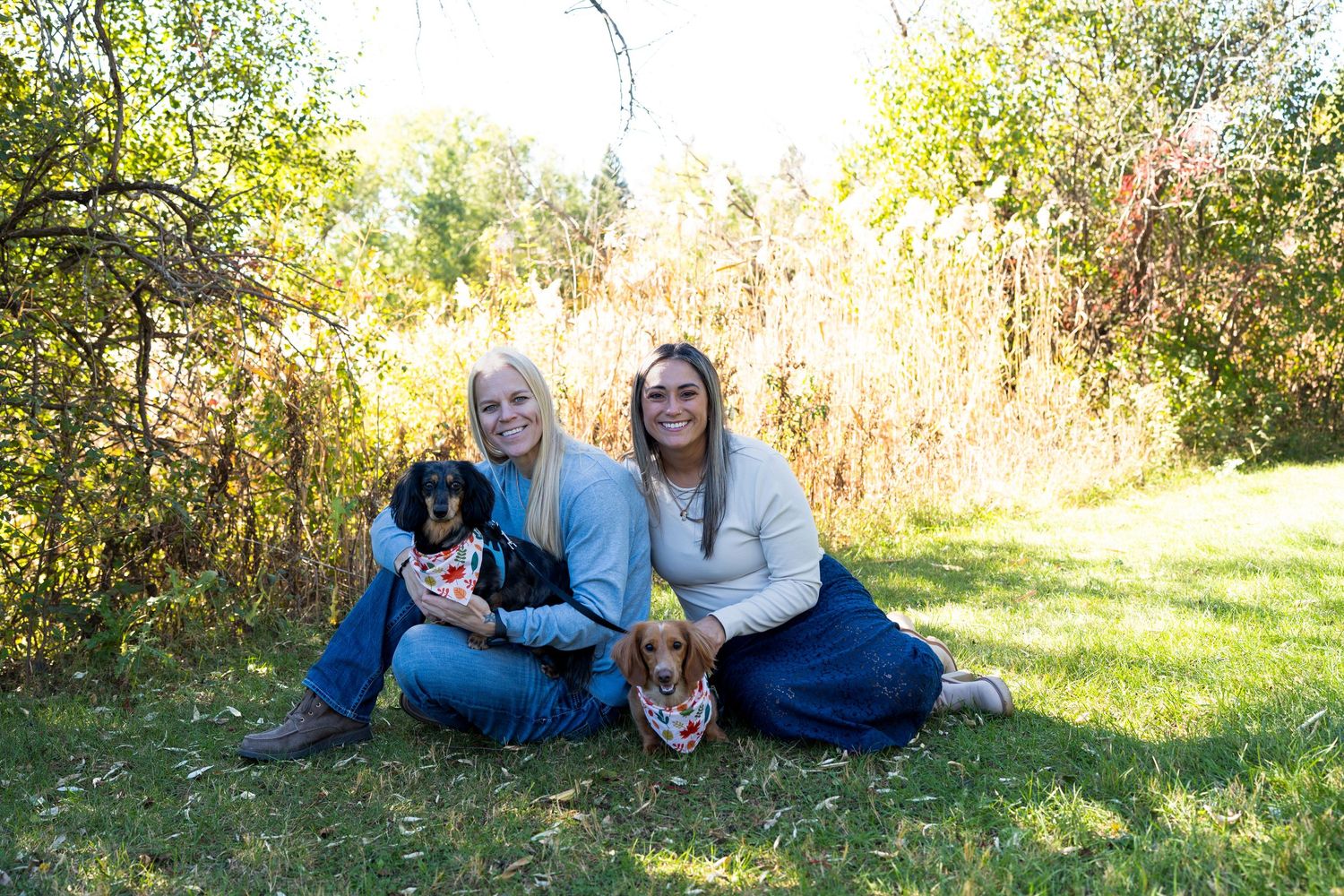 Two young women sit on grass with two dogs amid colorful autumn foliage.