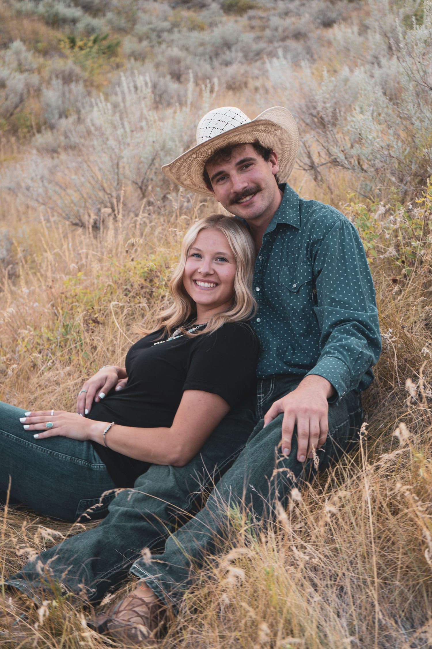 A smiling couple sits together in tall golden grass, with the man wearing a straw cowboy hat.