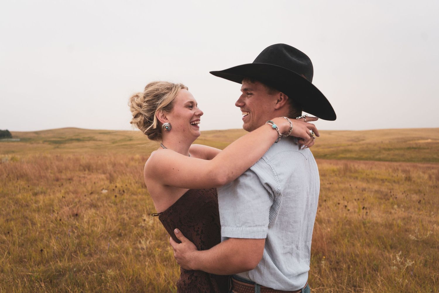 Newly engaged couple embracing and laughing in an open golden field, man wearing black cowboy hat.