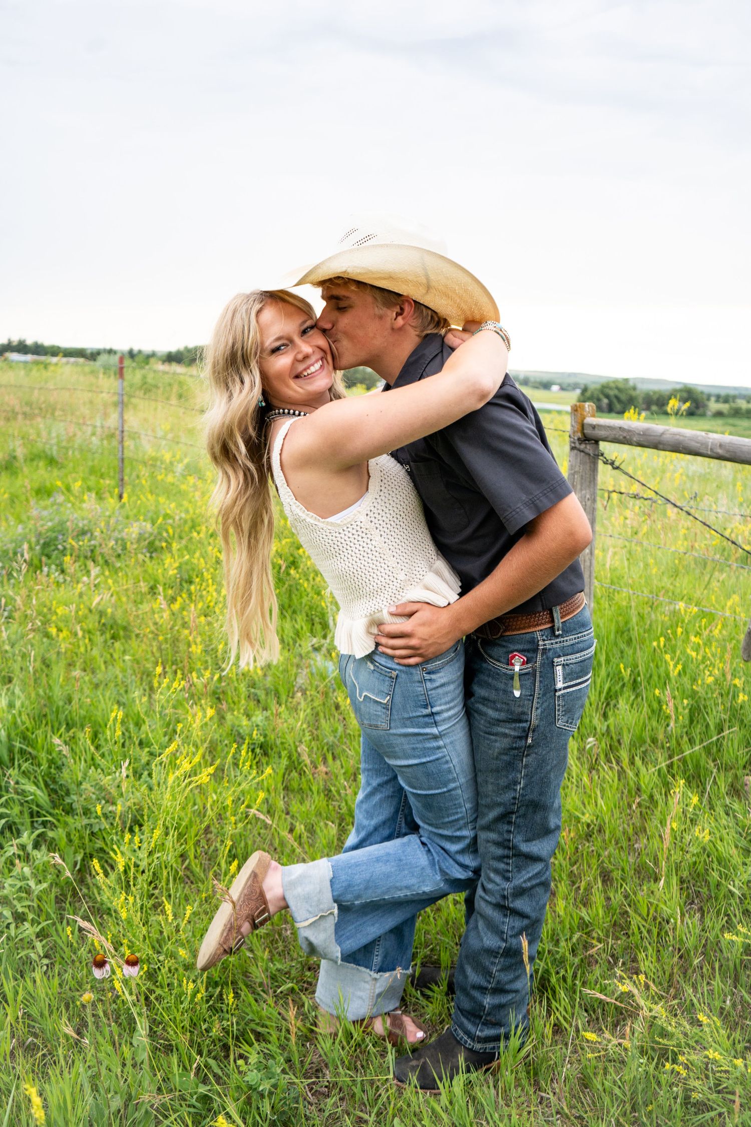 A cowboy kisses a smiling blonde woman in a green meadow near a fence.
