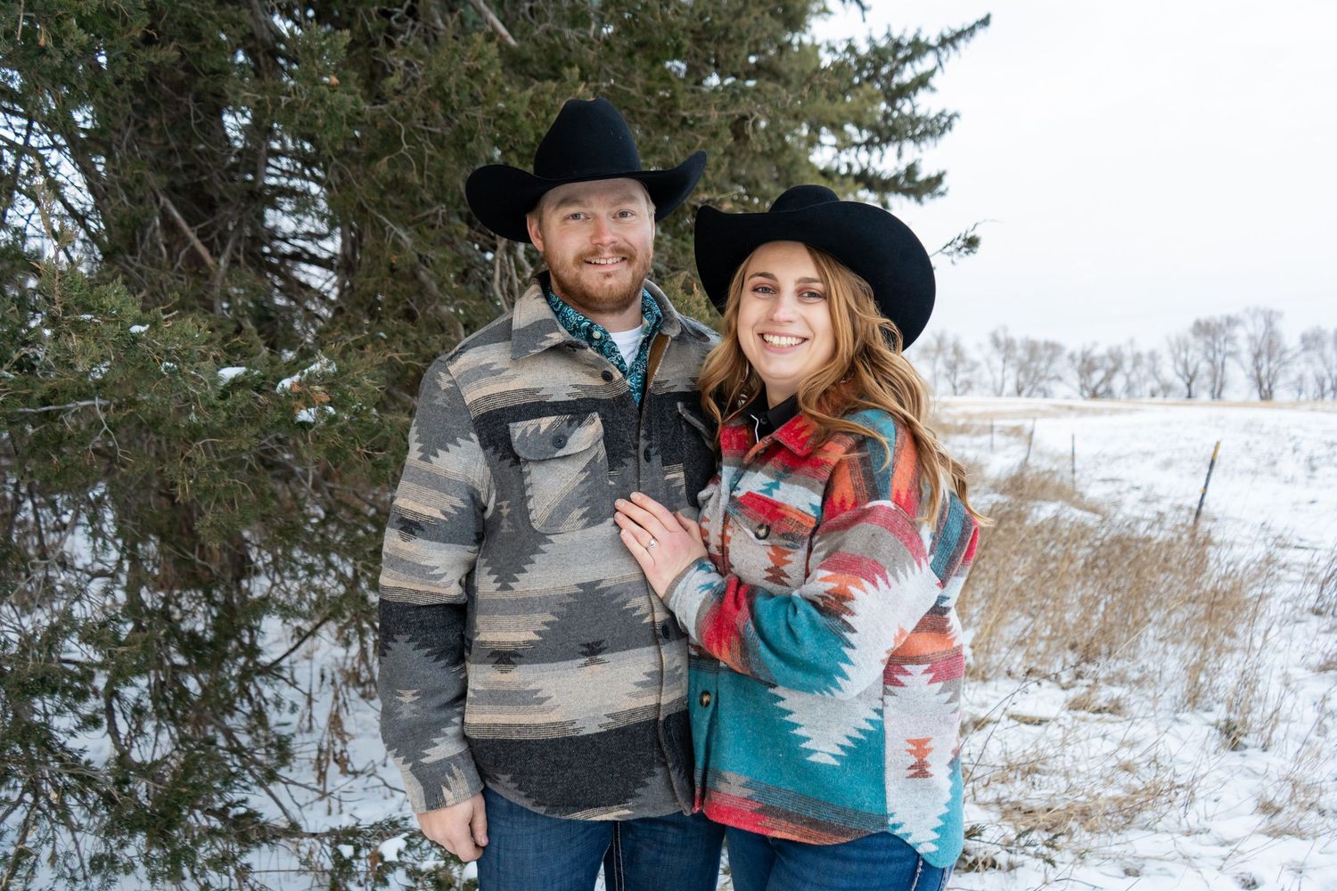 Smiling couple in Aztec print western jackets and black cowboy hats posing near a snowy pine tree.
