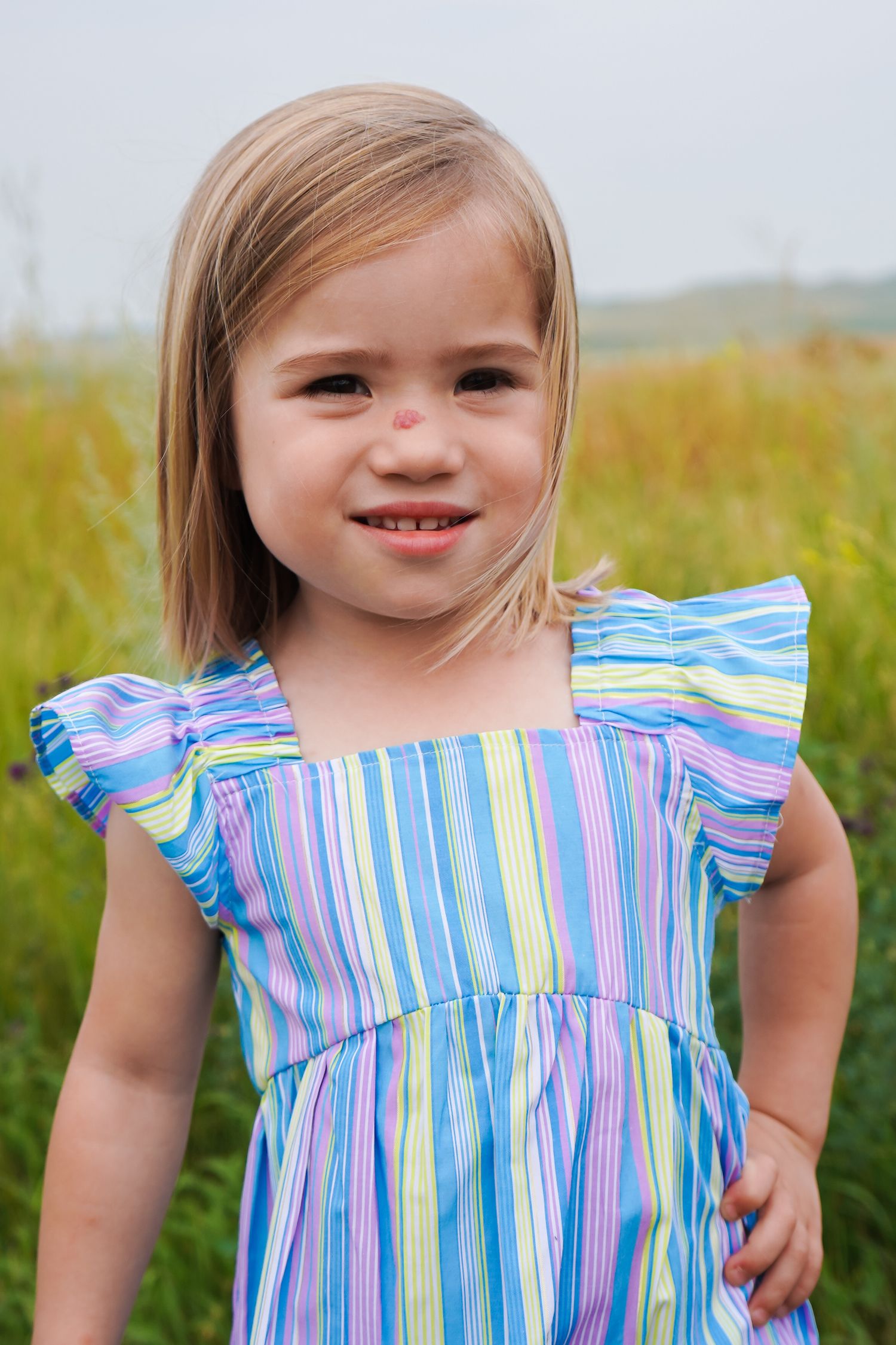 Cheerful blonde toddler girl wearing a multicolored striped dress standing in a grassy field.